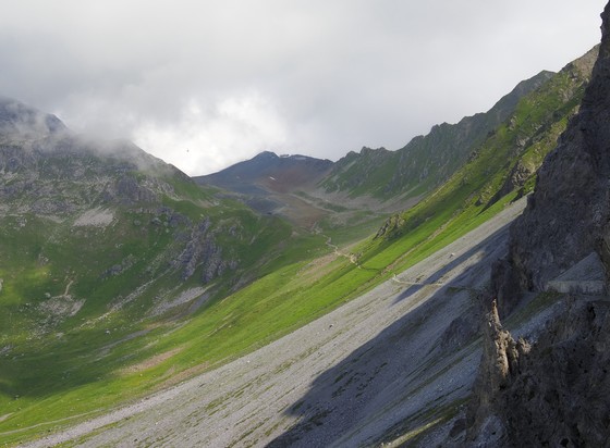 Mountain landscape with cloudy sky and lush green slopes, intersected by a winding path. In the distance a cable car descends through the clouds. 