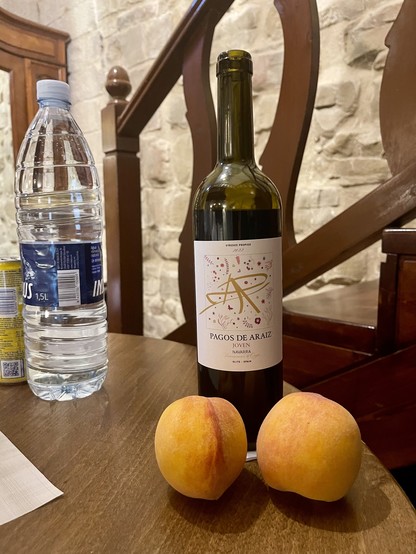 Eine Flasche Rotwein aus Olite/Navarra am Rande des runden Tisches, davor 2 typisch gelbe Weinbergspfirsiche, links davon eine Flasche Wasser, dahinter ein Treppengeländer aus Holz