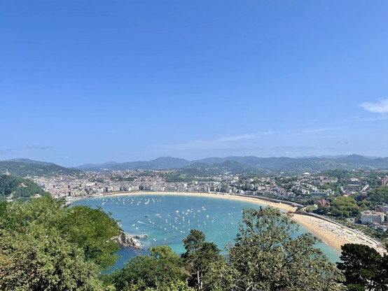 Bei schönstem Sonnenschein: die Concha aus der Höhe gesehen, Sandstrand und türkisblaues Wasser im vollendeten Halbkreis auf der rechten Seite, dahinter die Stadt und bewaldete Hügel, links Bäume und ein versteckter Blick auf Santa Clara.