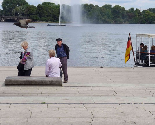 Ort: Binnenalster in Hamburg. Zwei ältere Personen begeben sich zu einer Steinbank. Eine Frau sitzt schon, die andere Frau ist kurz davor Platz zu nehmen. Ein älterer Herr wartet noch. Am linken oberen Fotorand fliegt eine Taube. Auf der rechten Seite des Fotos sieht man den hinteren Teil einer Barkasse mit ein paar Menschen und der Deutschlandfahne. Mittig im Foto die Wasserfontäne