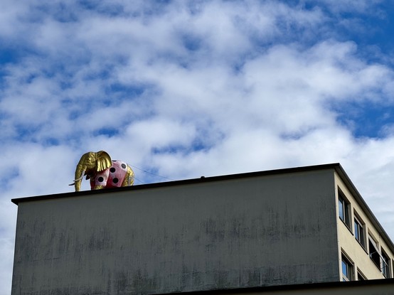 A large, colorful elephant sculpture with gold accents and polka dots is perched on the edge of a building roof, set against a backdrop of blue sky and clouds.