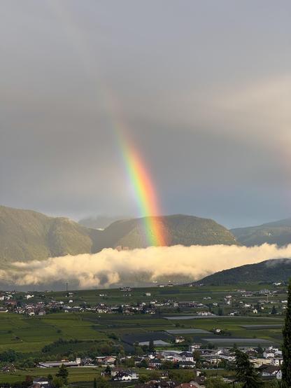 A vibrant rainbow arcs over green hills and valleys, with clouds and mist settling among the landscape and a small town visible below. The scene captures a serene, post-rain atmosphere.