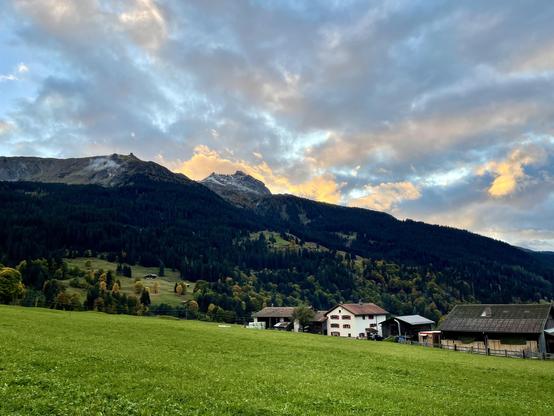 Bild mit Bergpanorama, Herbstwäldern und grünen Wiesen.