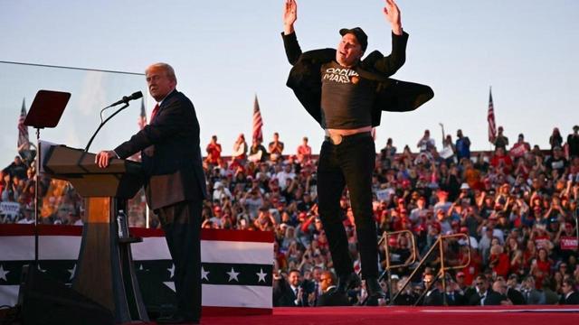 A rally scene featuring a weird senile speaker at a podium, with a crowd visible in the background. A weird super-rich person jumps enthusiastically nearby, wearing a black jacket and a graphic t-shirt stating his maniac dream. Flags and banners are present, indicating a political event for massive idiots. 