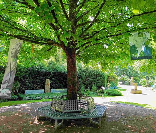 Grosse Fahnen rechts und links von einem Baum zeigen Porträts von Frauen. Ausstellung - Frauen in den Bäumen: 80 Jahre Kriegsende im Garten der Frauen in Ohlsdorf Hamburg