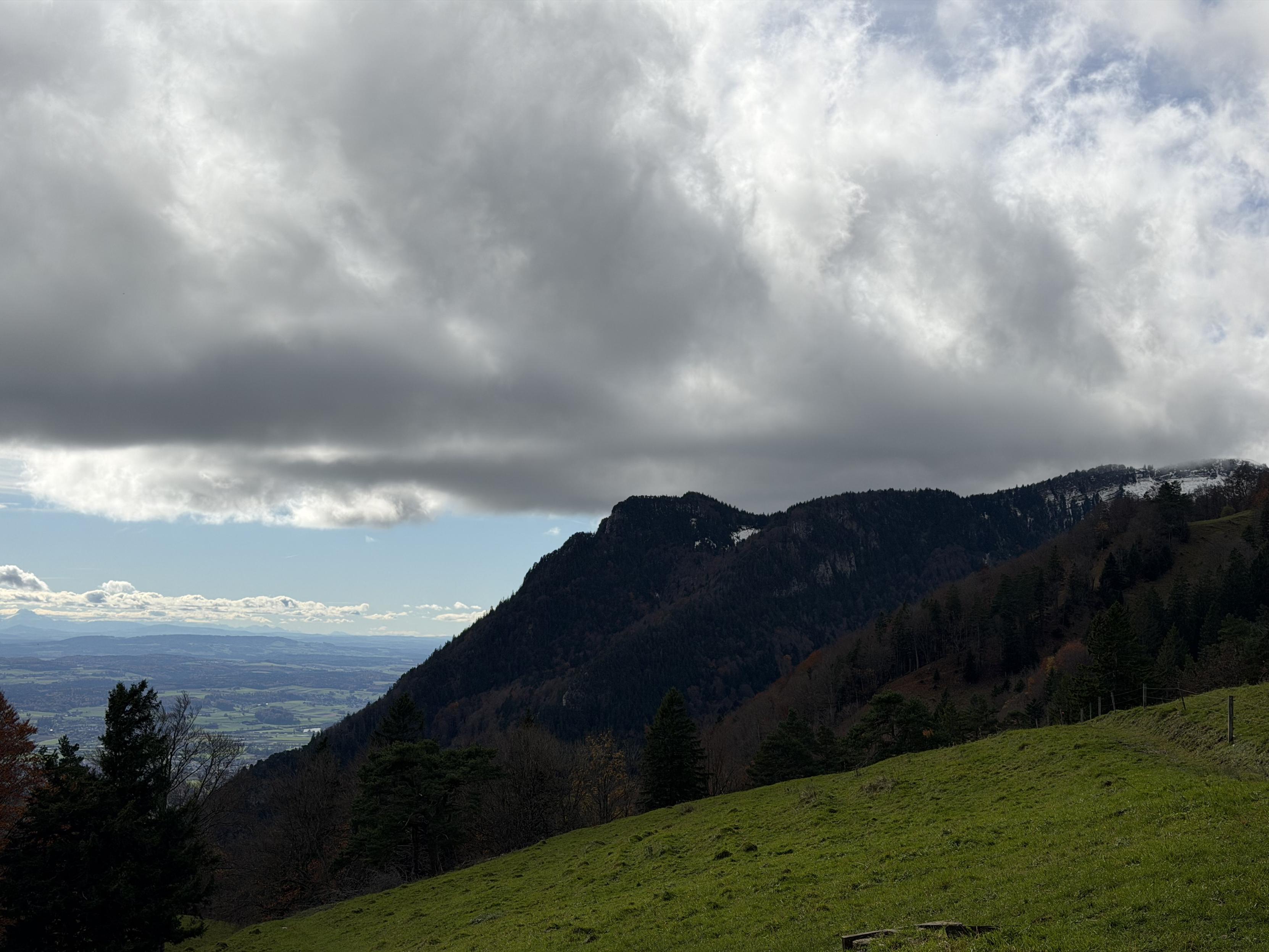 Graue Wolken mit etwas blauen Himmel, in der Mitte Berge welche leicht mit Schnee bedeckt sind, links die Sicht ins Tal. vorne Bäume und Wiese
Grey clouds with some blue sky, mountains lightly covered with snow in the middle of the picture, view of the valley to the left. Trees and meadow in the foreground.