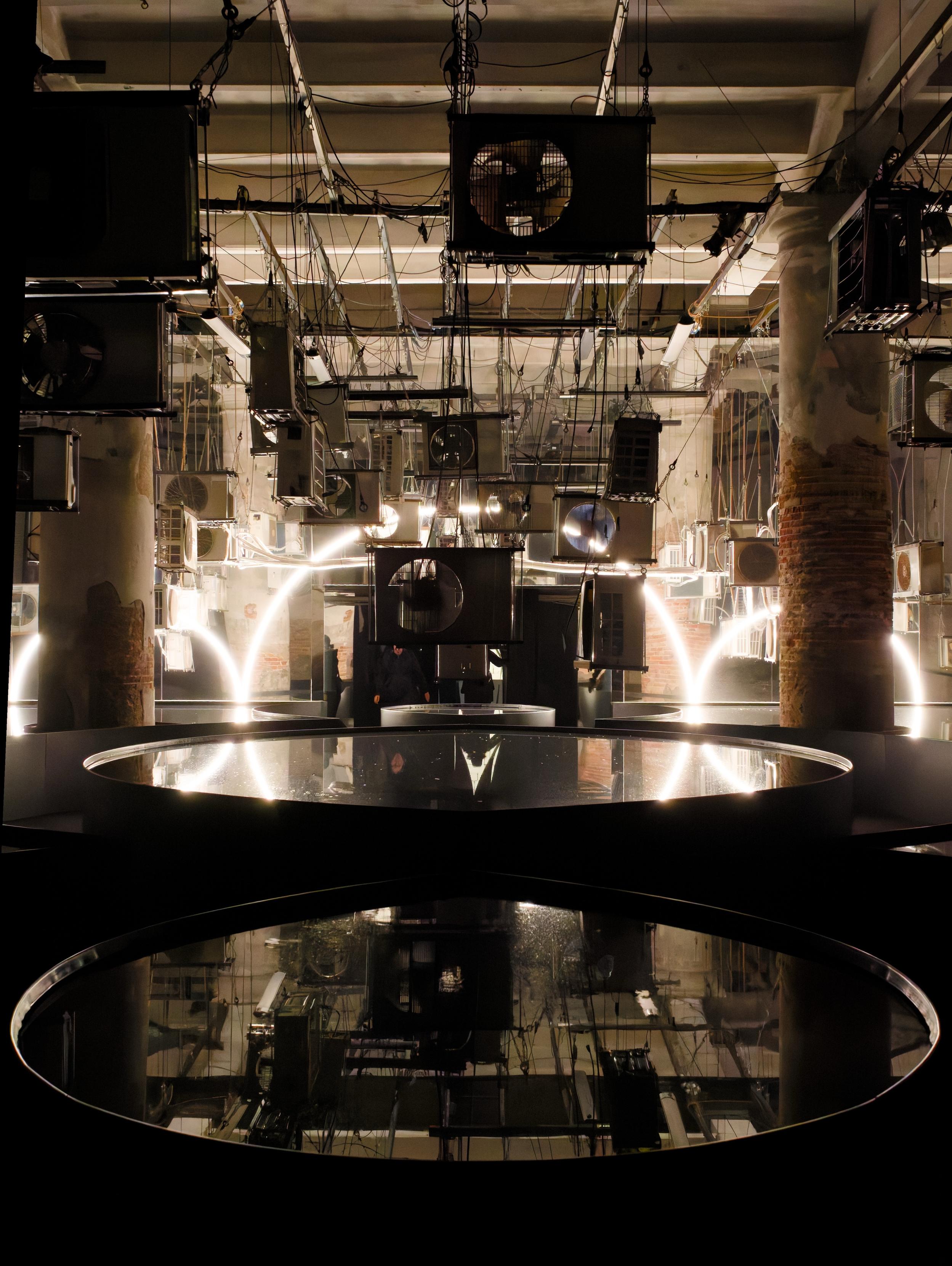 An industrial space featuring numerous air conditioning units and intricate wiring suspended from the ceiling. Two circular reflective surfaces are positioned in the foreground, capturing the ambient light and surroundings. The environment has a dimly lit, artistic atmosphere.