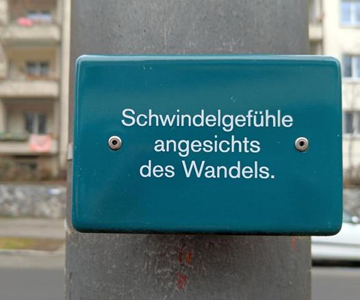 Kleines Schild angebracht an einer Strassenlaterne mit der Aufschrift: Schwindelgefühle Angesicht des Wandels