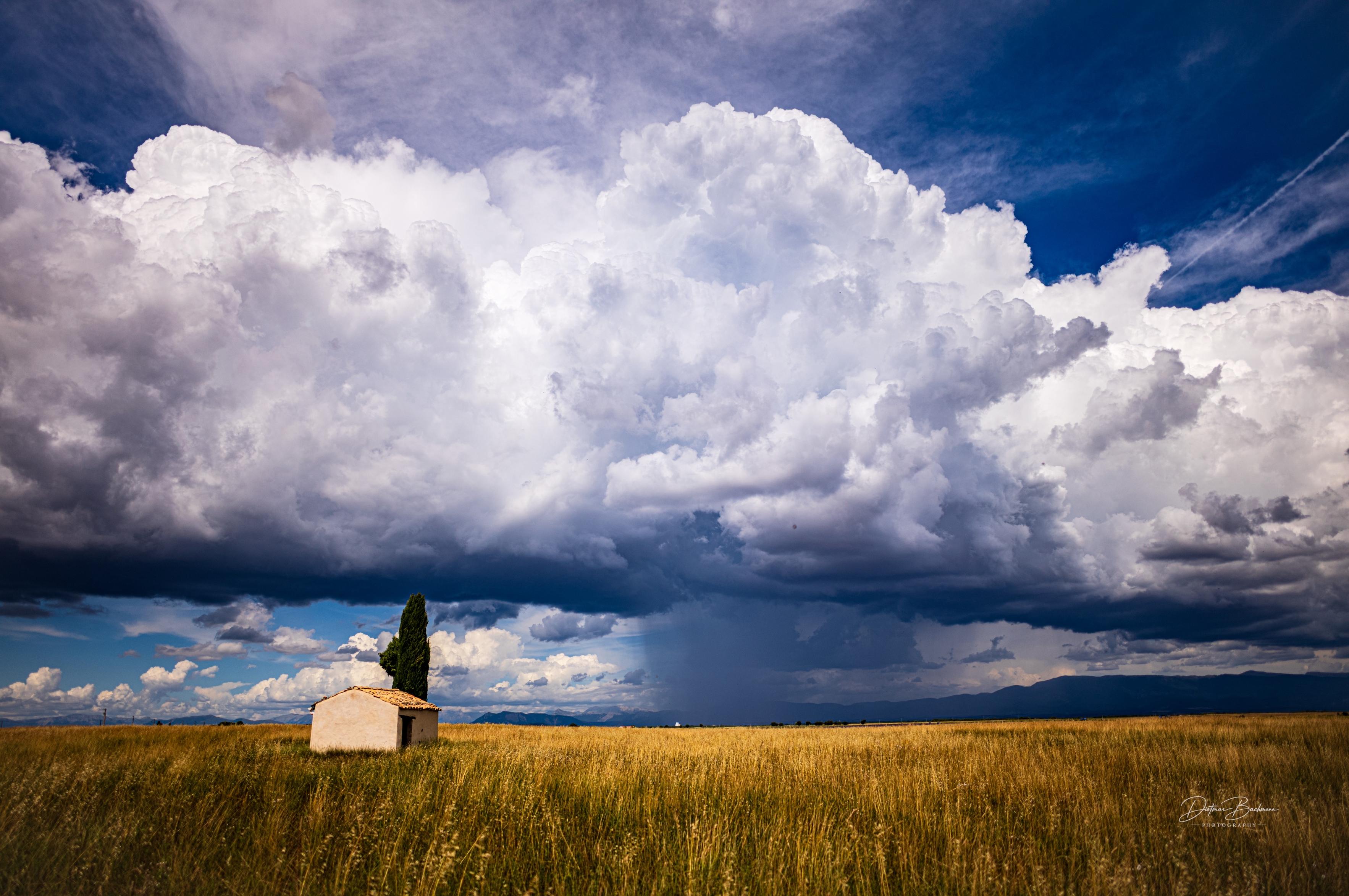 A solitary white building sits in a golden field under a dramatic sky filled with large, fluffy clouds, some darker, indicating possible rain. A tall tree is visible near the structure, with distant mountains in the background.