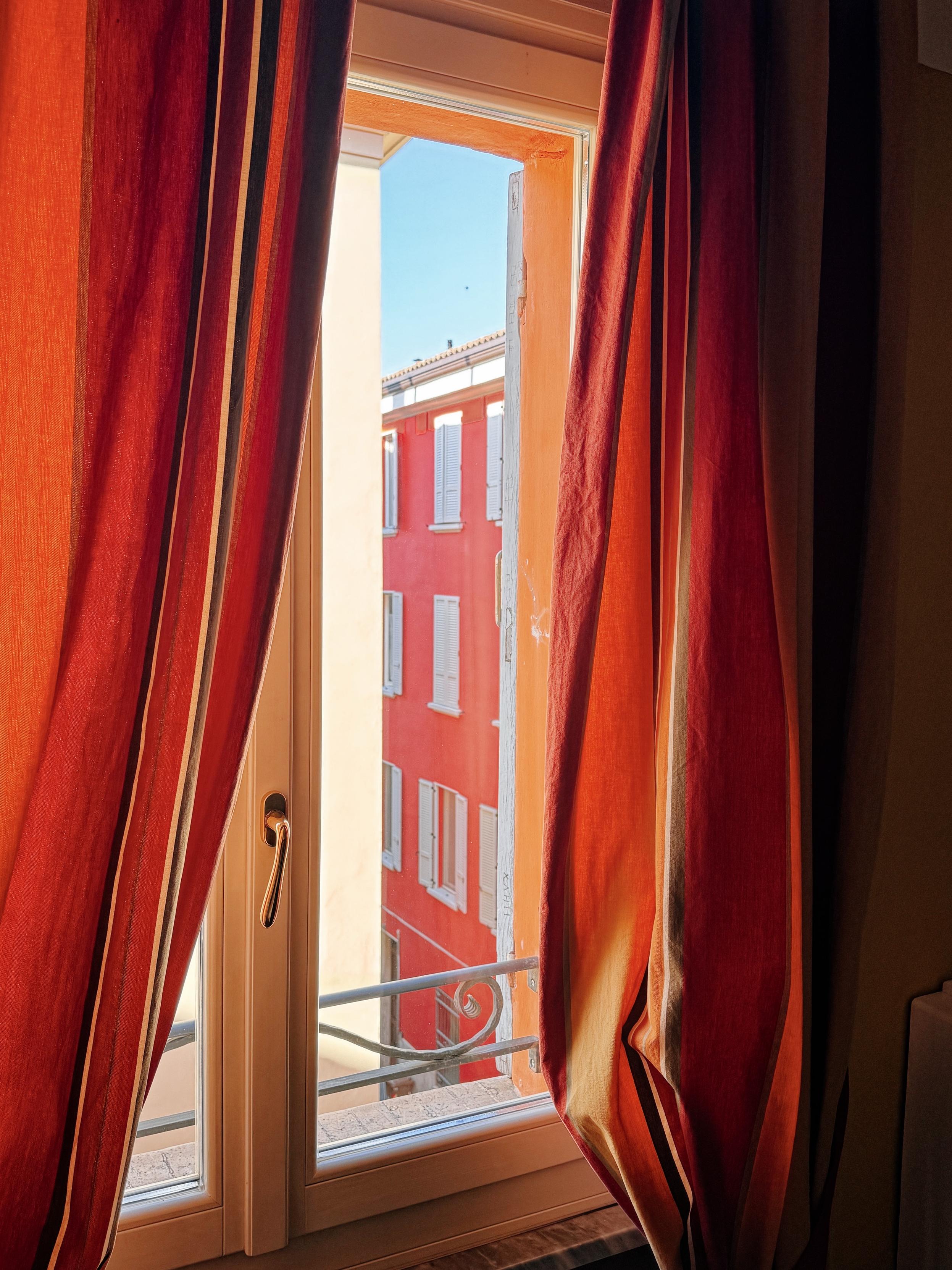 A window framed by red and orange curtains reveals a view of a red building across the street and a clear blue sky.