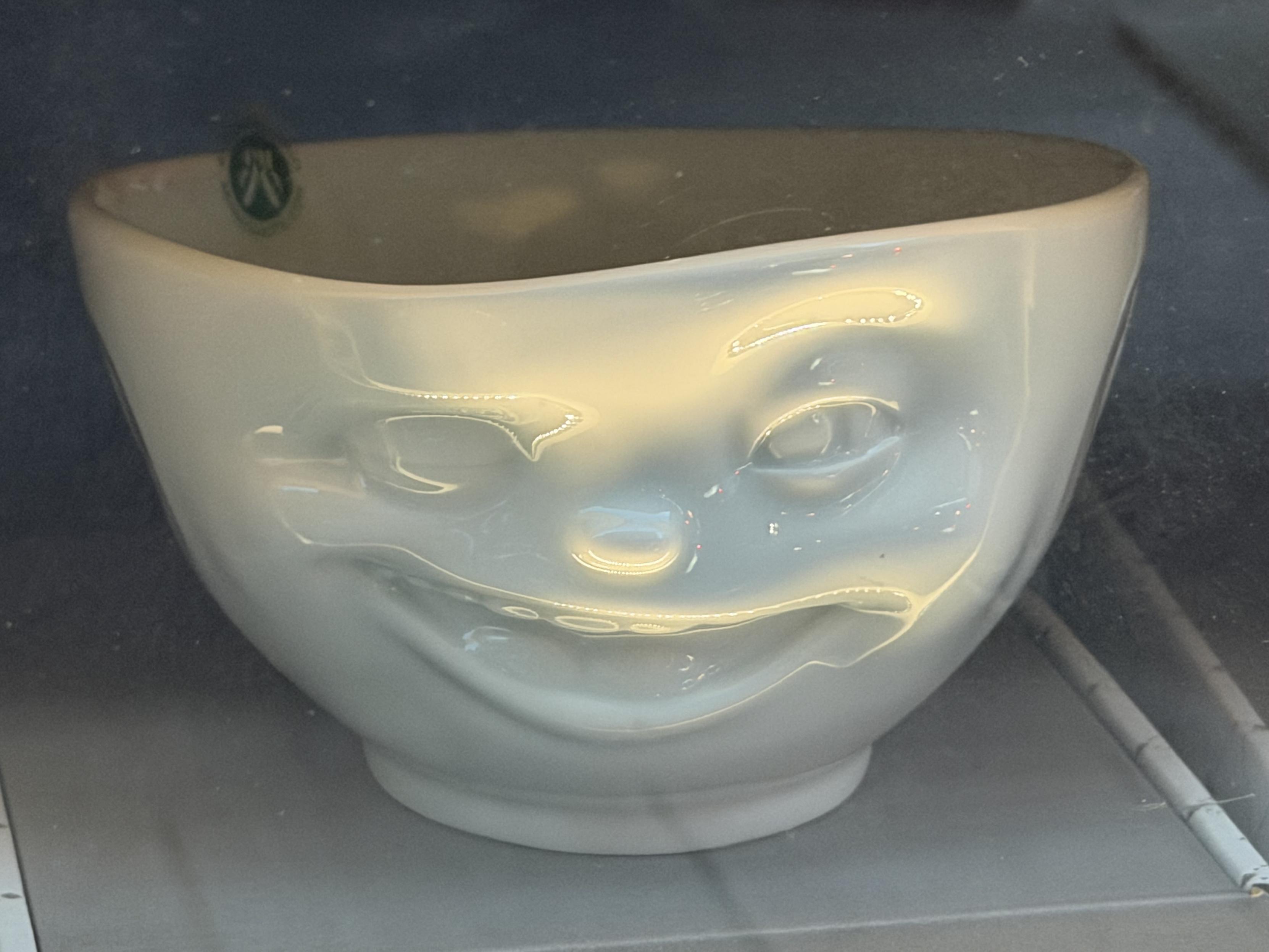 A white bowl in a shop window. It has an evil grinning face on it and is illuminated in a way that looks scary.
