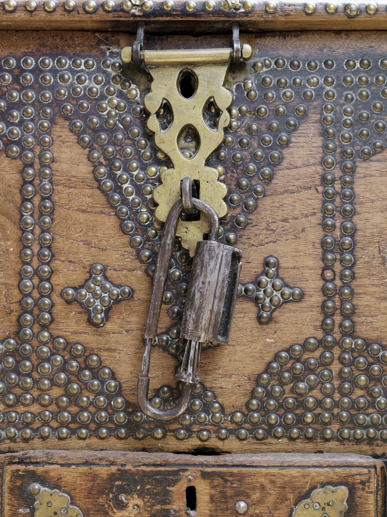 An ornate wooden chest showing decorative brass hardware, including a lock and hinges, complemented by an intricate pattern of brass studs.