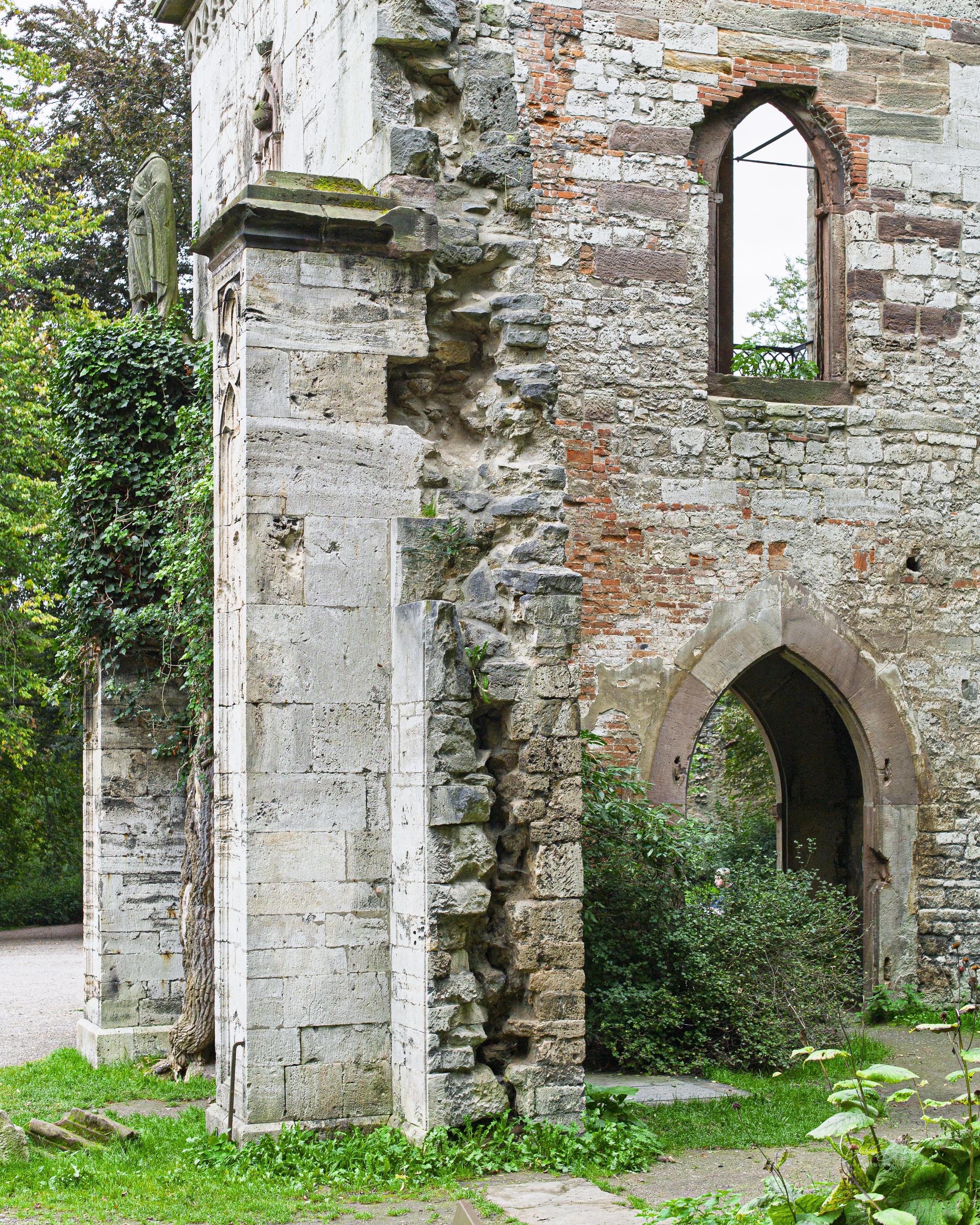 A close-up of a weathered stone wall featuring a mix of brick and rough stone textures, partially covered with greenery. An old window with an arch shape is visible. The scene conveys a sense of age and nature reclaiming the structure.
