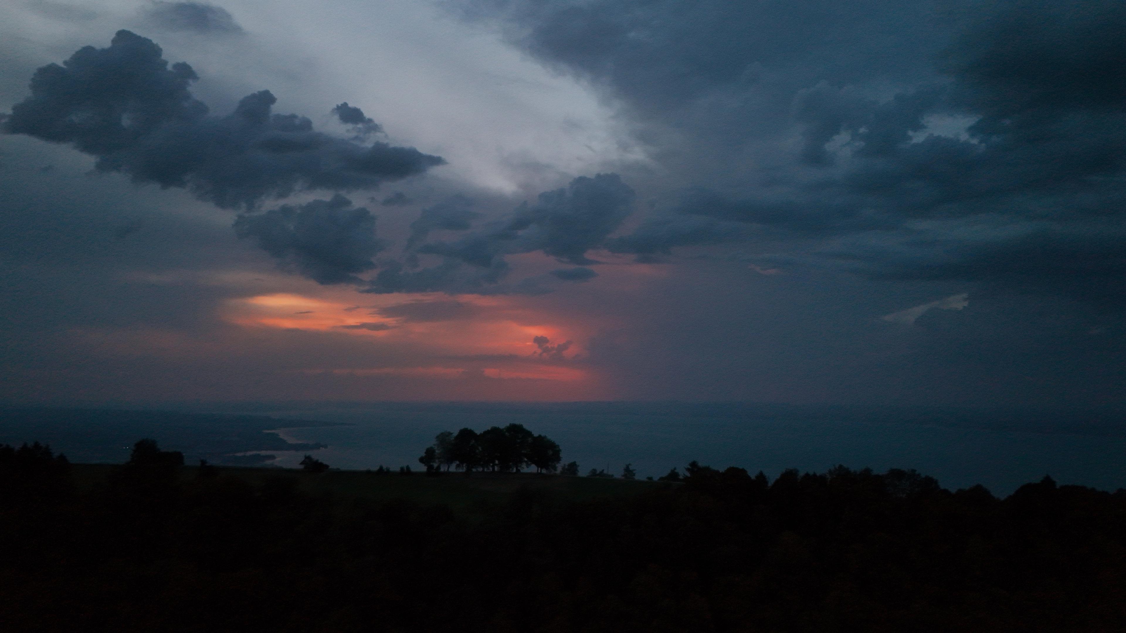 Ein dramatischer Sonnenuntergang über einem Gewässer mit dunklen Wolken und einem Hauch von orangefarbenem und rosa Licht am Horizont. Der Vordergrund zeigt Silhouetten von Bäumen vor einem sich verdunkelnden Himmel.