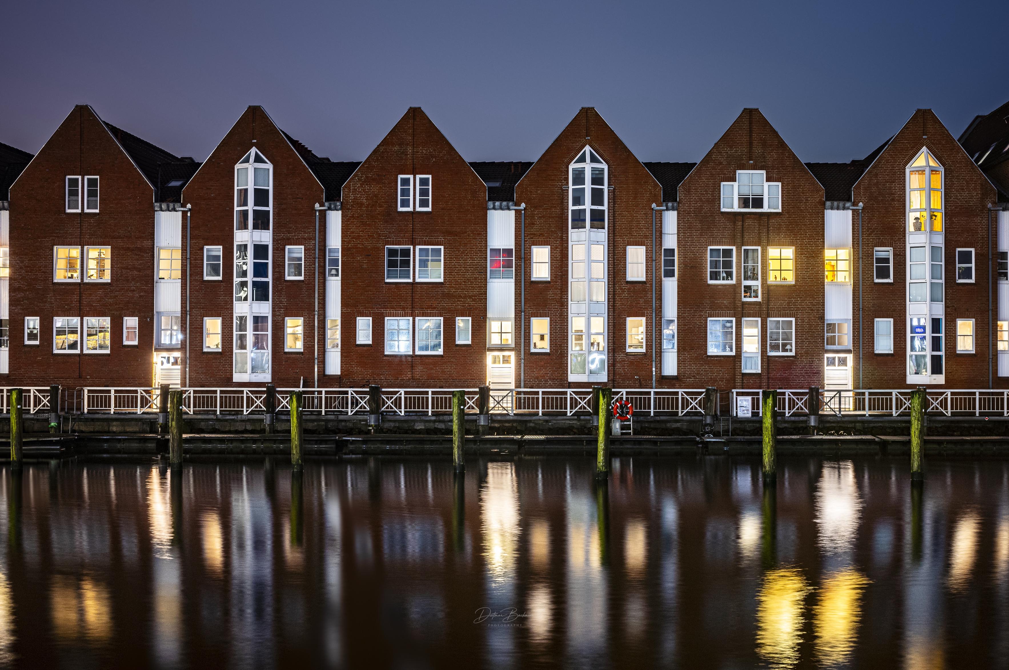 A row of modern brick buildings with slanted roofs, reflecting in calm water at night. Illuminated windows suggest activity inside, while a dock with wooden posts is visible in the foreground. The overall atmosphere is serene and urban.
