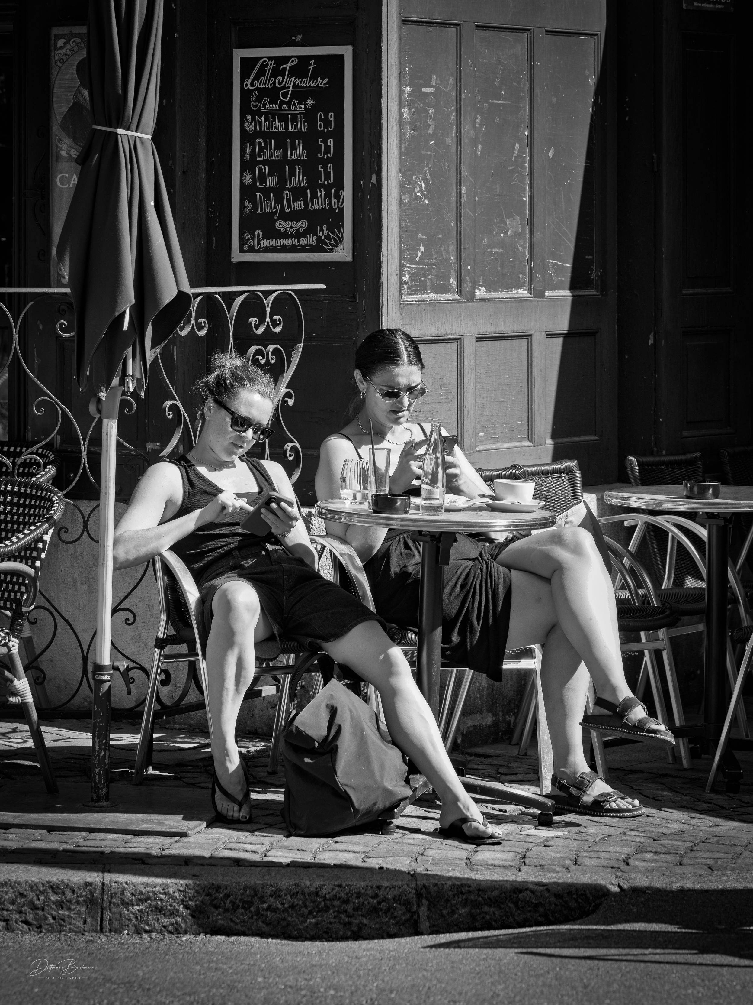 Two women sit at an outdoor café, absorbed in their phones. They wear sunglasses and casual black dresses. In the background, there's a chalkboard menu displaying various latte options. The scene is in black and white, emphasizing a relaxed, candid atmosphere.