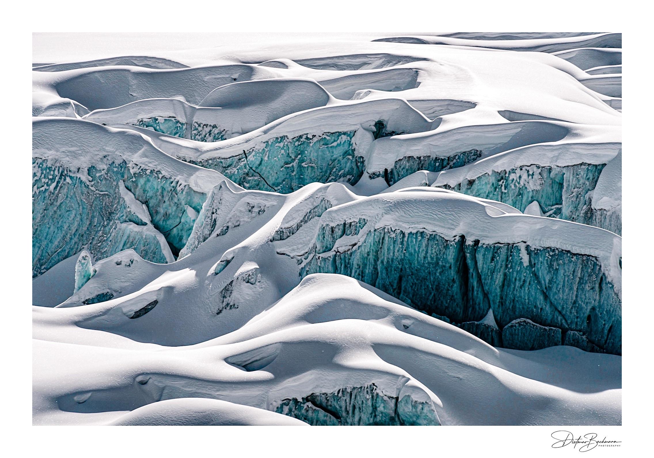 An aerial view of a glacier landscape featuring smooth, white snowdrifts and deep, turquoise-blue crevices. The contrasting colors and textures create a striking, icy terrain.