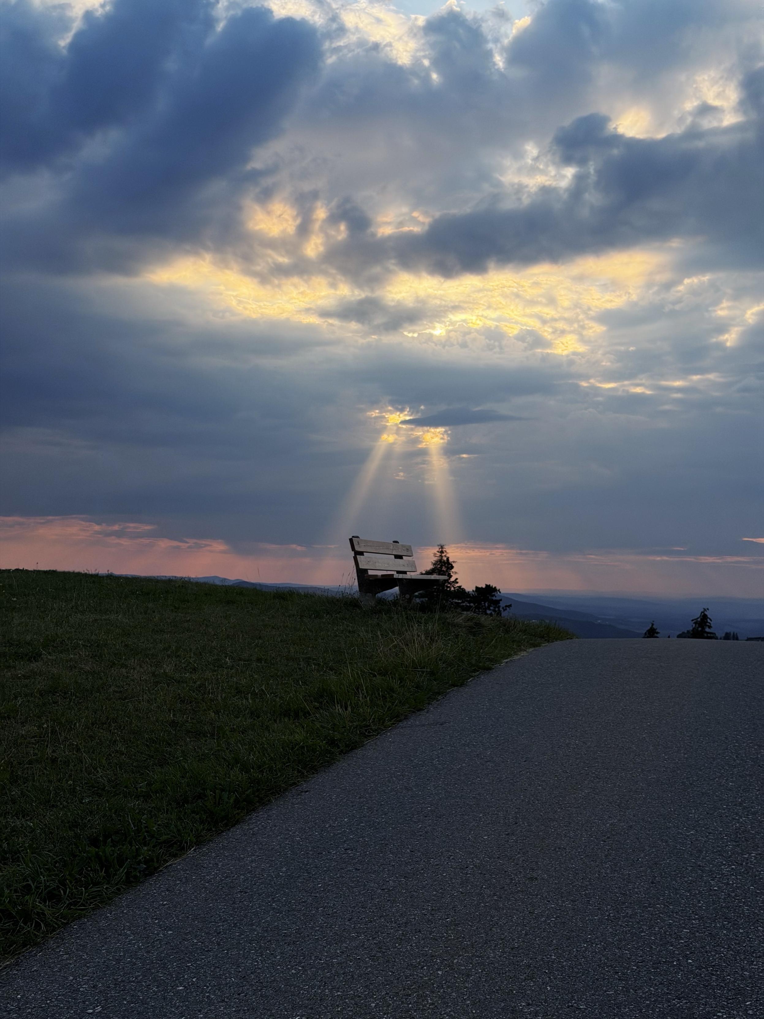 Eine Holzbank auf einem grasbewachsenen Hügel überblickt einen gewundenen Weg, mit dramatischen Wolken und Sonnenstrahlen, die bei Sonnenuntergang durch den Himmel brechen und eine ruhige Landschaft schaffen.