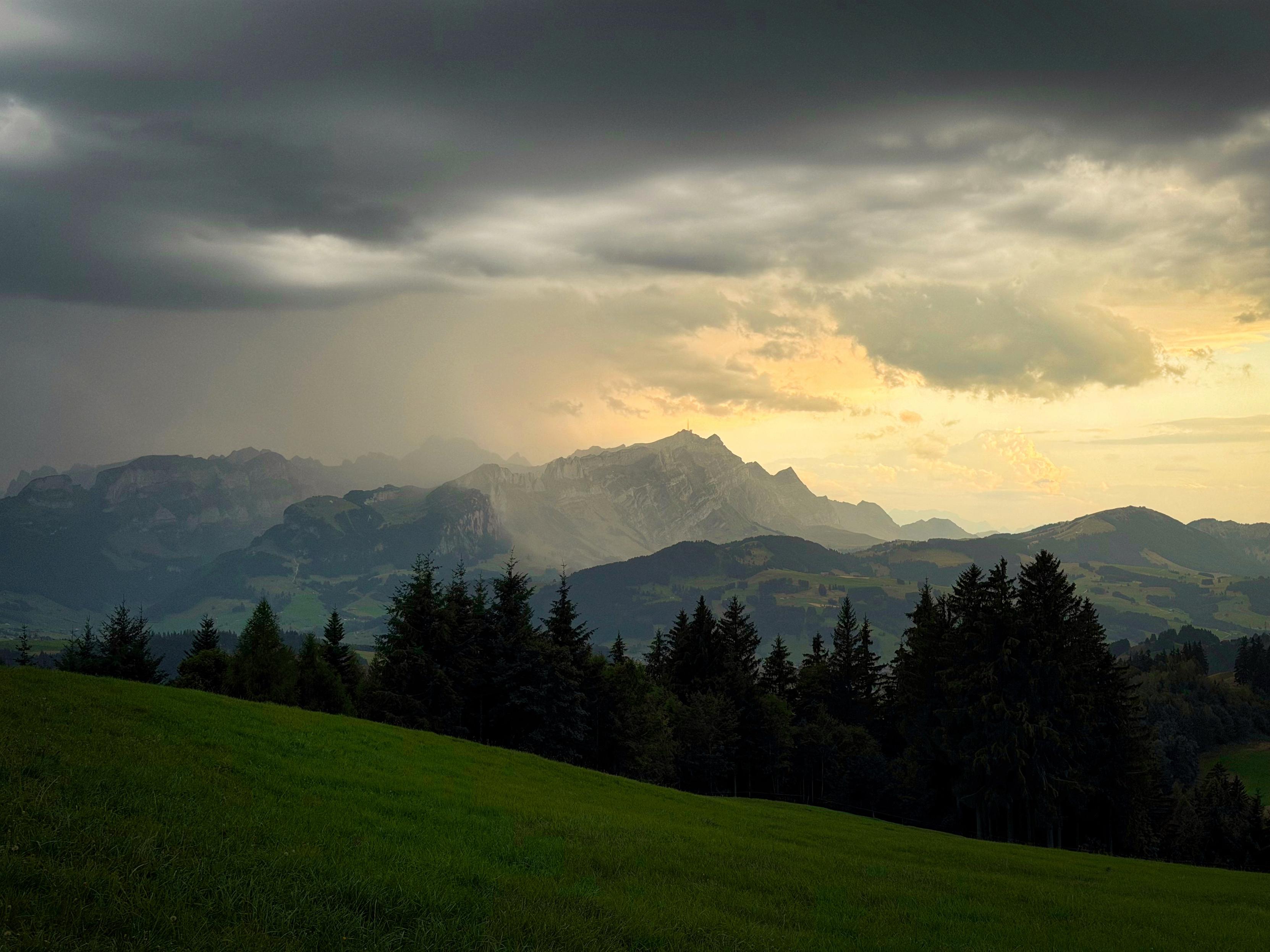 Ein Panoramablick auf eine Berglandschaft unter einem bewölkten Himmel, mit dunklen Wolken und einem Hauch von Sonnenuntergang im Hintergrund. Grünes Gras und Nadelbäume sind im Vordergrund sichtbar, im Kontrast zu den zerklüfteten Bergen in der Ferne.