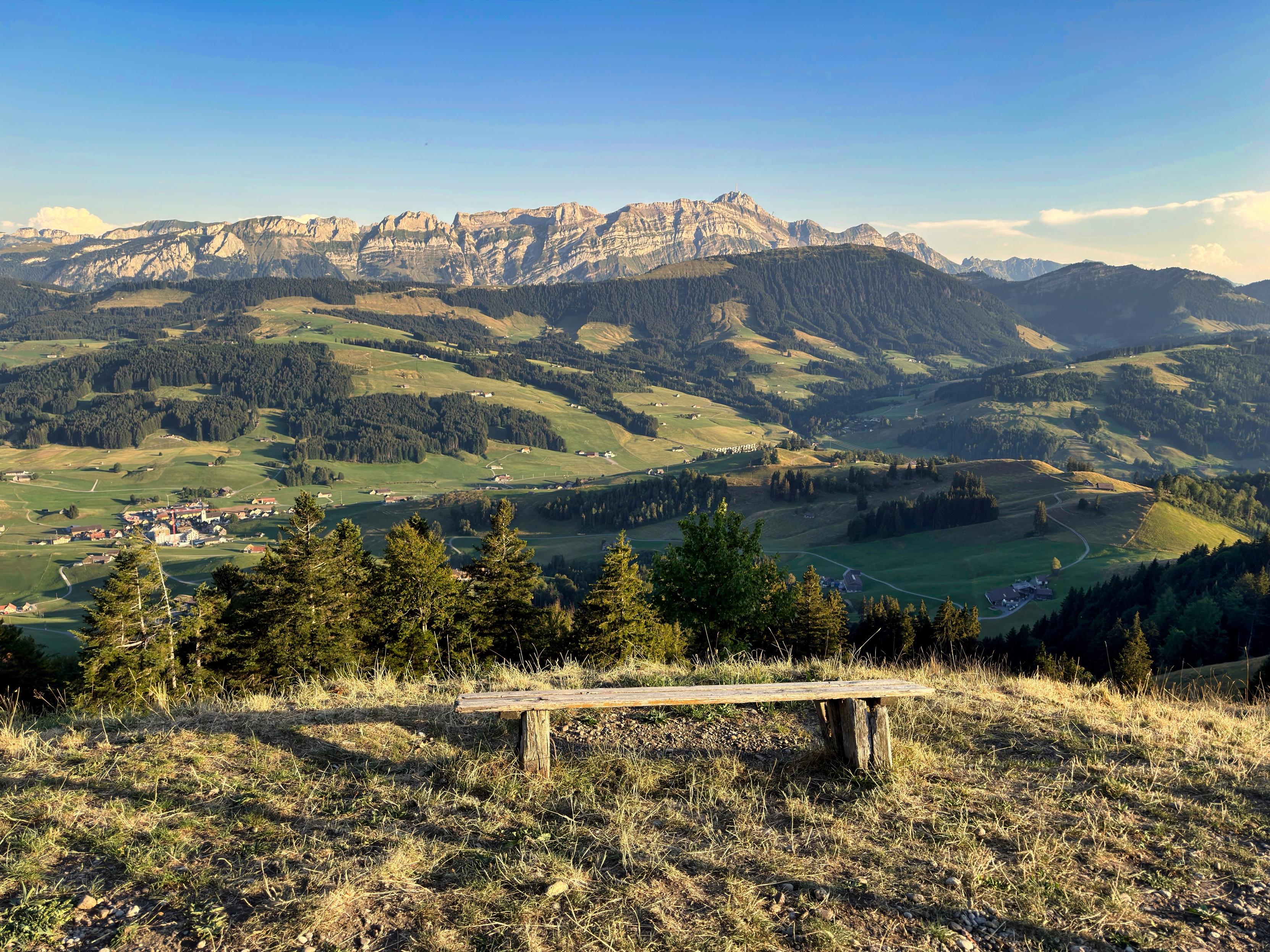 Ein Panoramablick auf eine Berglandschaft mit sanften Hügeln und einem kleinen Dorf im darunter liegenden Tal. Im Vordergrund steht eine Holzbank auf einer grasbewachsenen Fläche mit Blick auf die Landschaft, mit Bäumen, die die Aussicht umrahmen. Der Himmel ist klar.