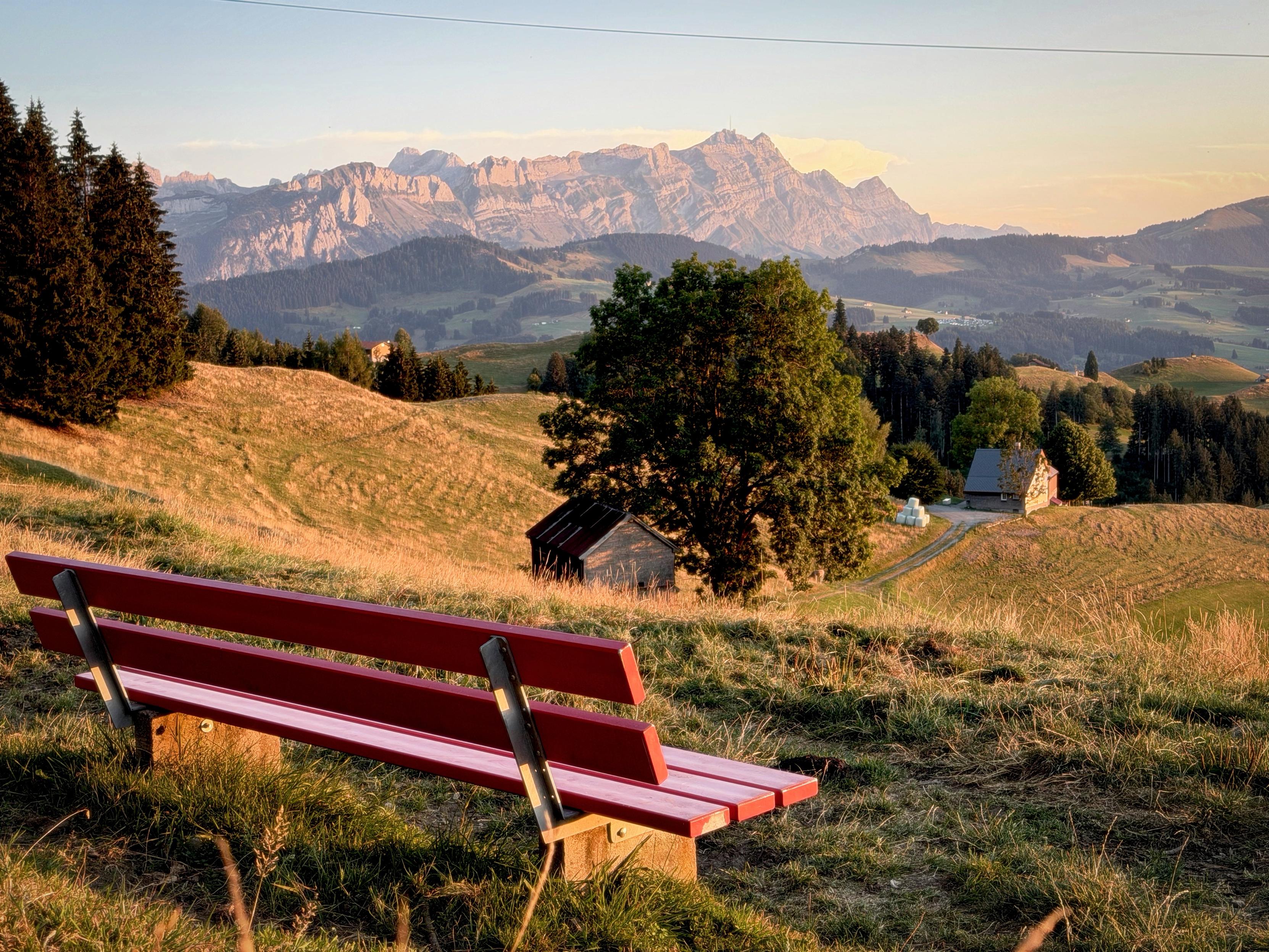 Eine rote Bank überblickt eine malerische Landschaft mit sanften Hügeln, Bäumen und fernen Bergen. Die untergehende Sonne wirft ein warmes Leuchten.