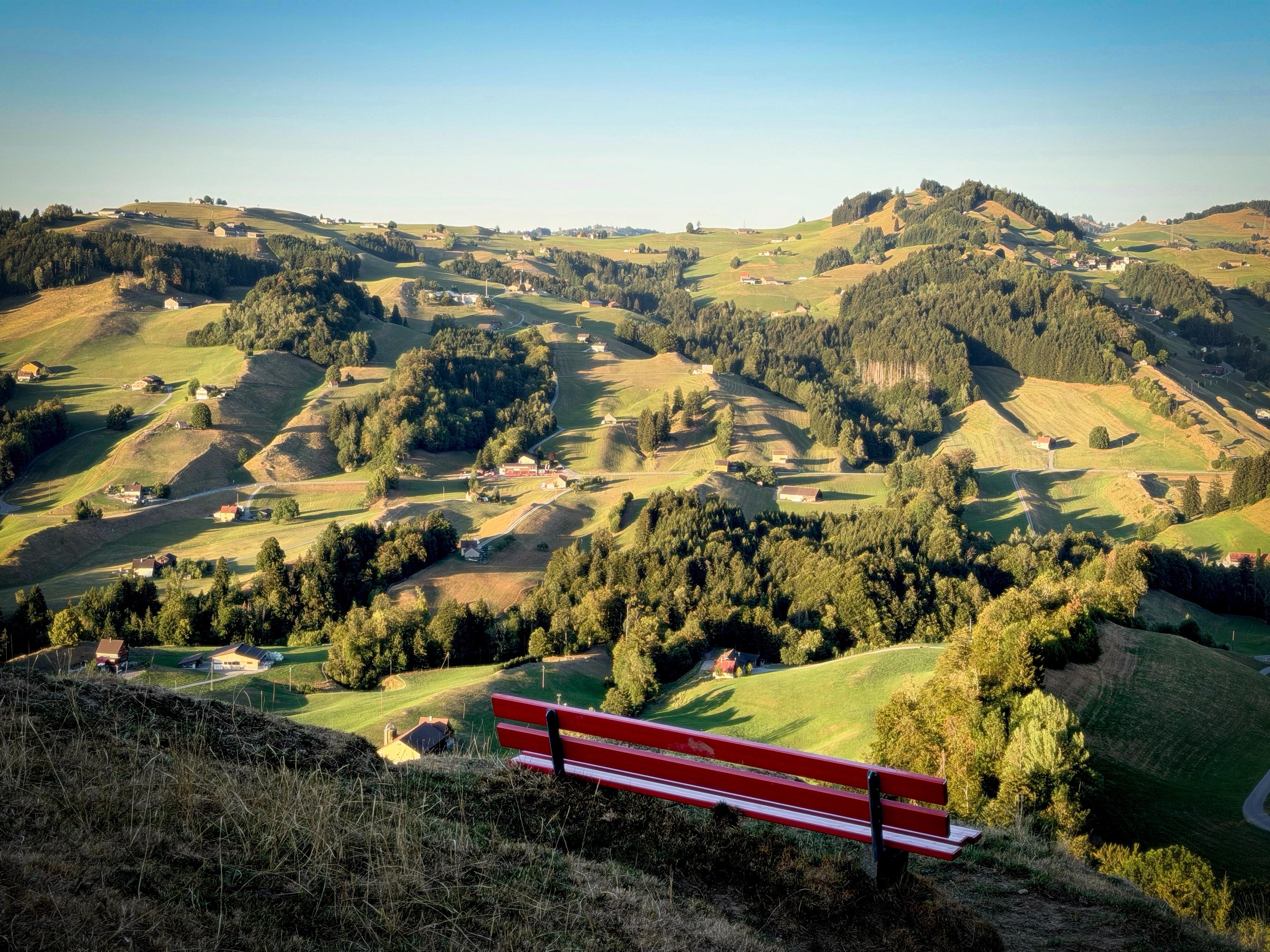 Eine malerische Landschaft mit sanften grünen Hügeln und Tälern, übersät mit kleinen Häusern und Bäumen. Eine rote Bank steht im Vordergrund und lädt zur Betrachtung der ruhigen Aussicht ein. Der Himmel ist klar und hell, was die ruhige Atmosphäre verstärkt.