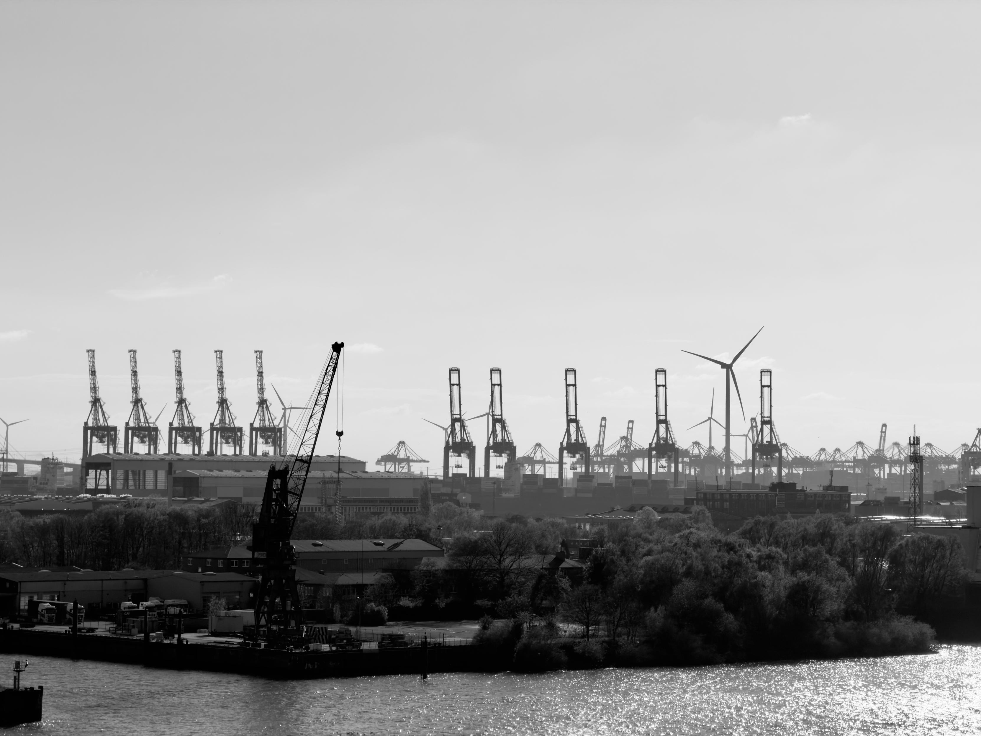 Schwarz-weißes Hafenpanorama mit Kränen, Windrädern, Containern, Bäumen und einem Kran im Vordergrund am Wasser.