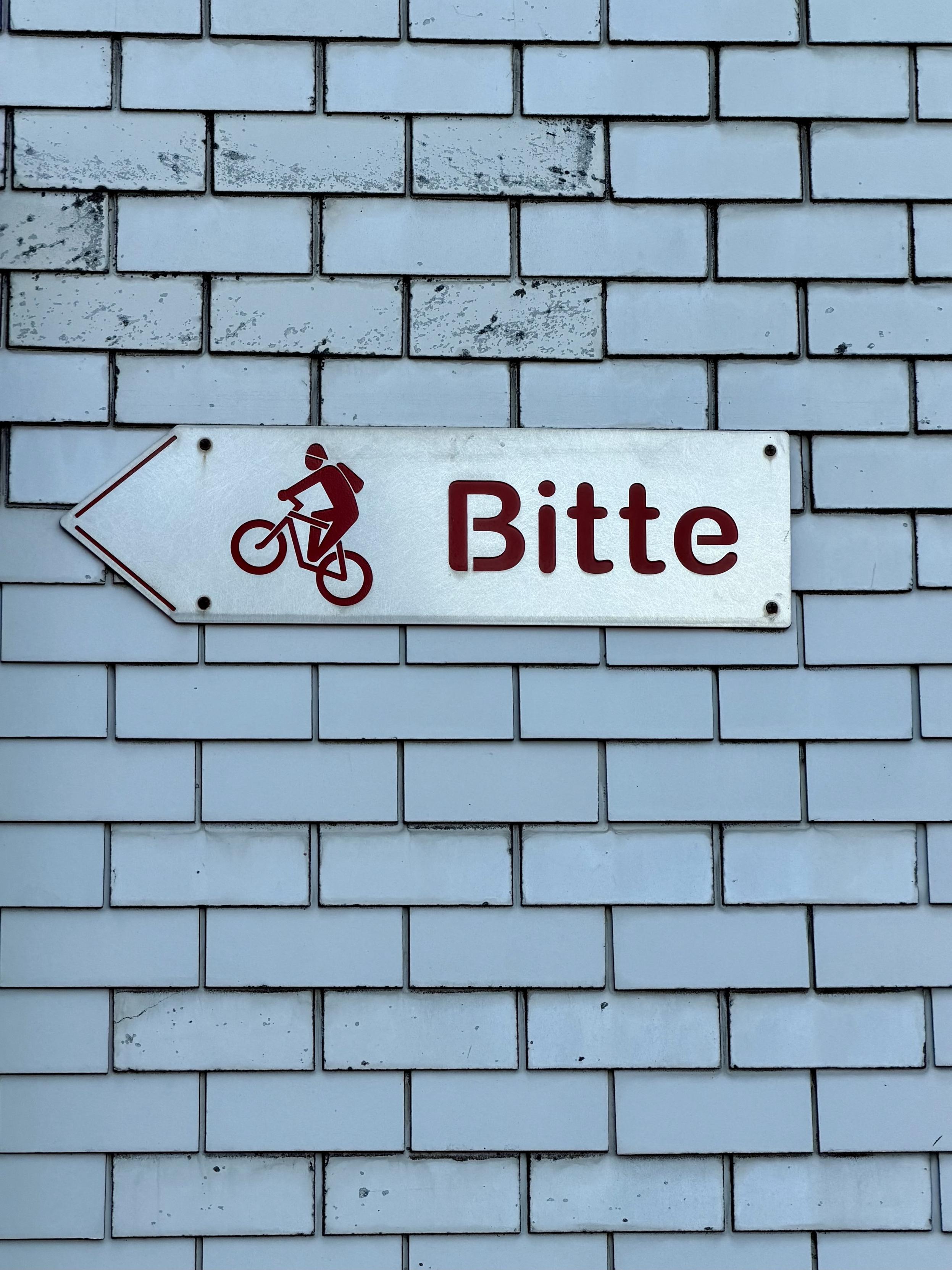 A street sign indicating a bike path, featuring an illustration of a cyclist and the word "Bitte" (meaning "please" in German). The sign is mounted on a light blue brick wall.