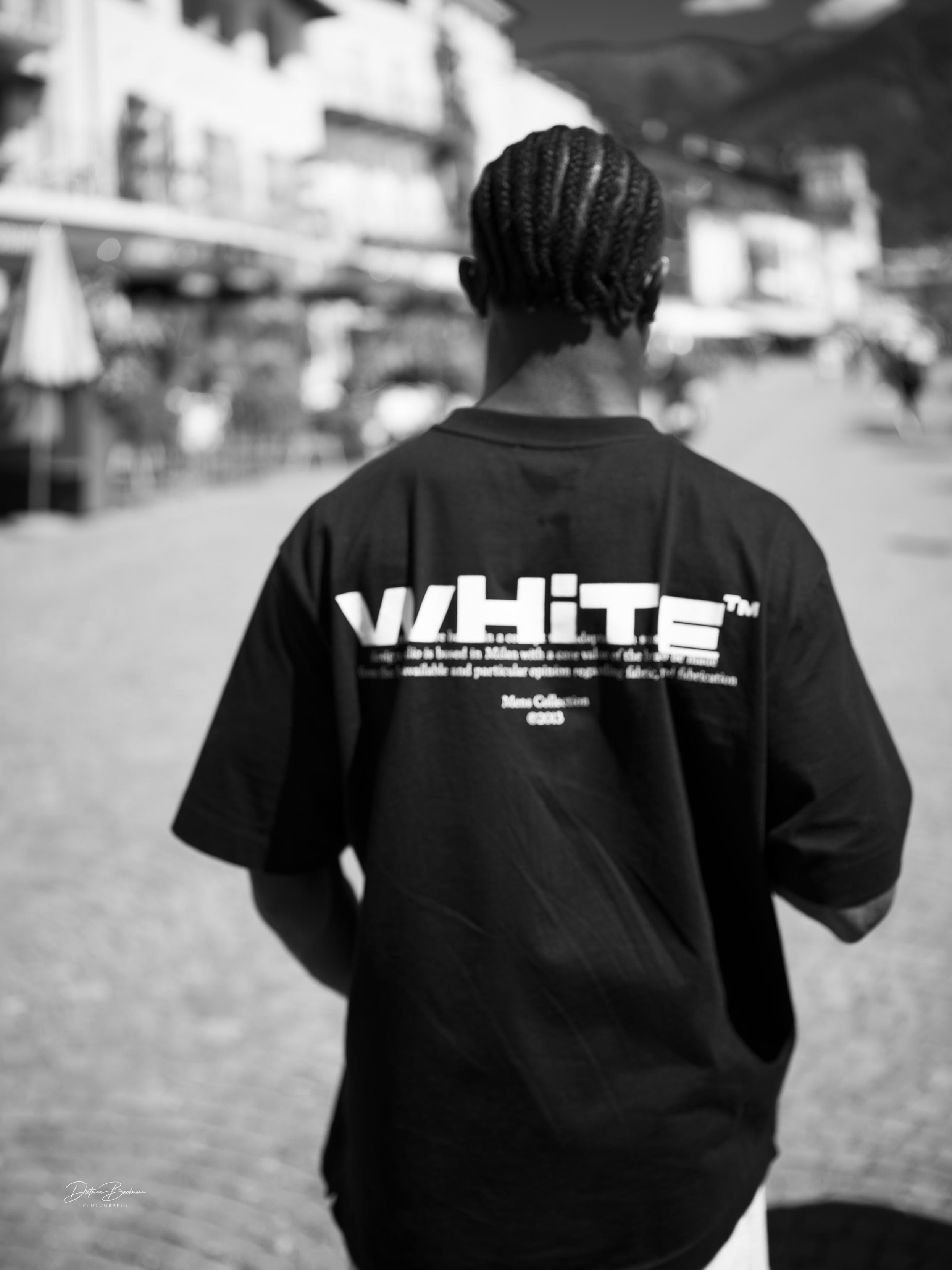 A person with braided hair is seen from the back, wearing a black t-shirt with the word "WHITE" prominently displayed. The background features a blurred street scene with buildings and people, captured in black and white.
