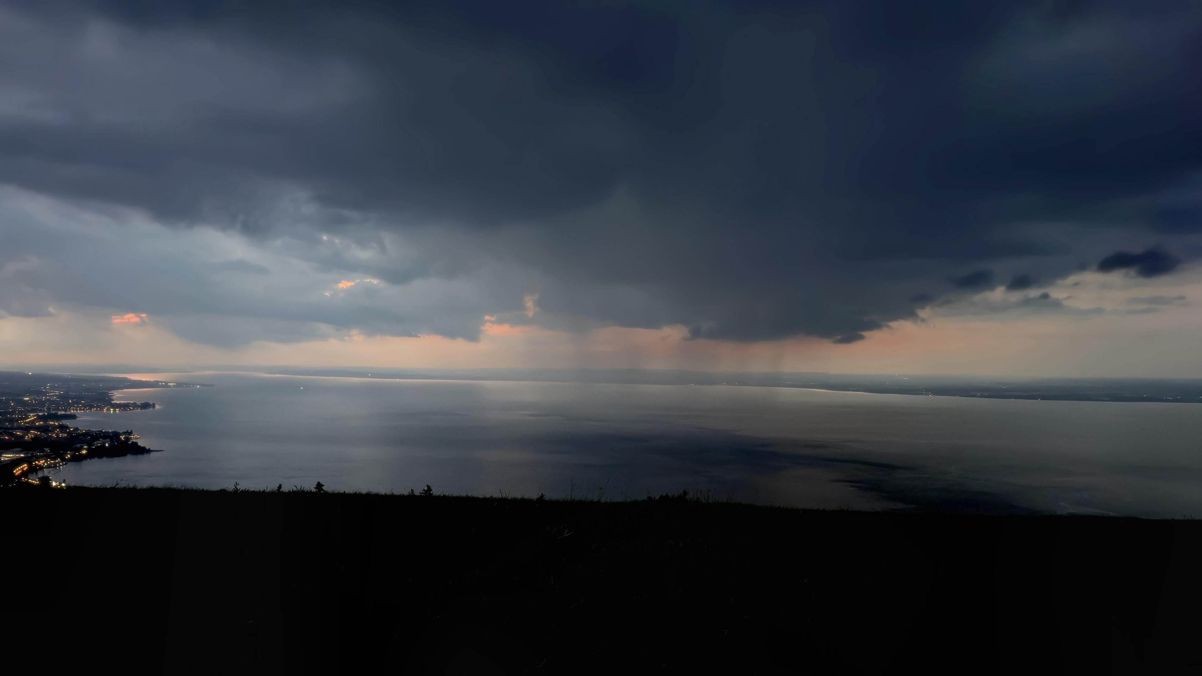 Ein Panoramablick auf einen dunklen, stimmungsvollen Himmel über einem ruhigen Gewässer. Der Horizont zeigt einen Hauch von Rosa und Orange, während der Vordergrund entfernte Stadtlichter entlang der Küste zeigt.