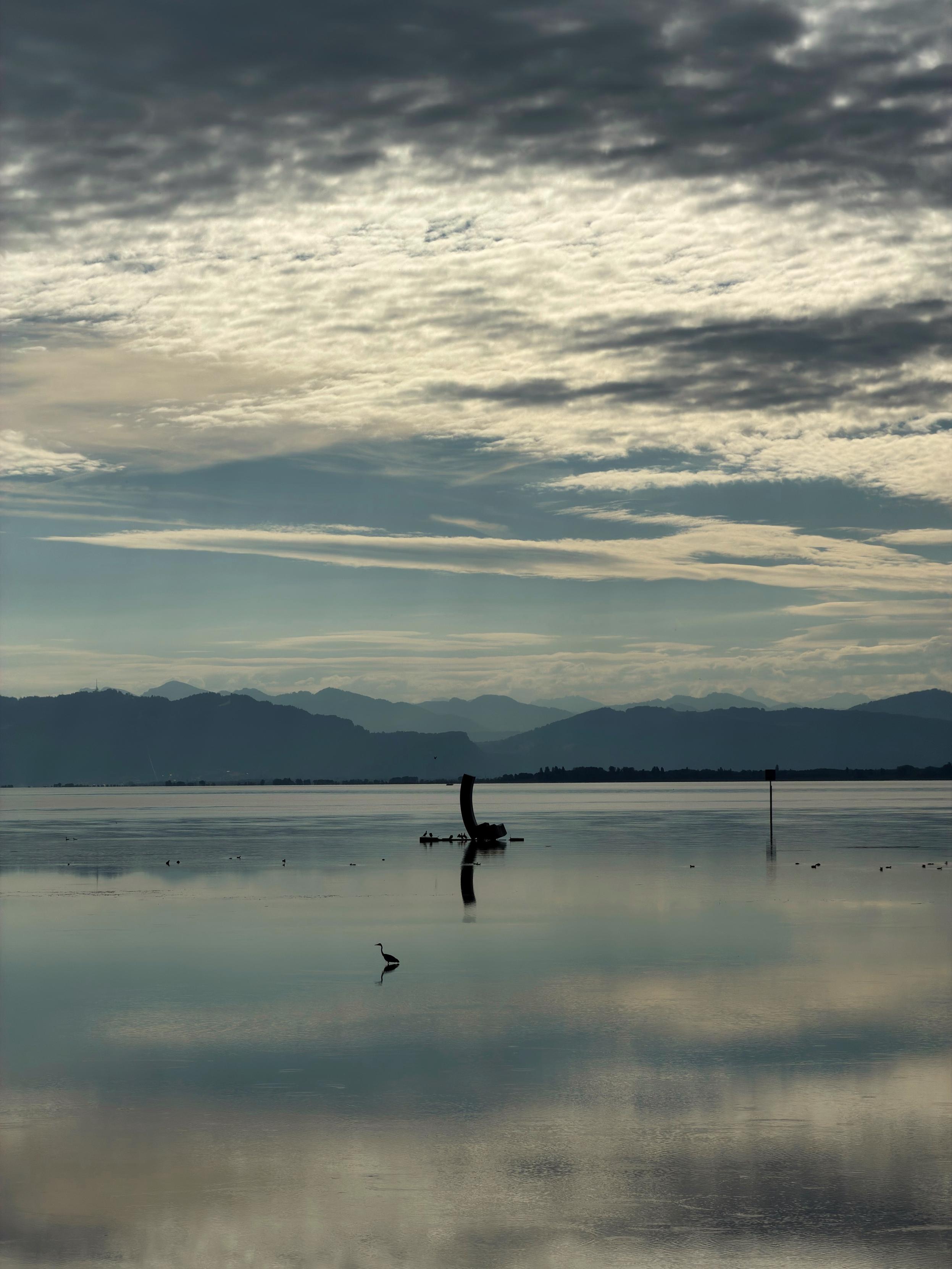 Eine ruhige Seeszene im Morgengrauen, mit ruhigem Wasser, das ferne Berge und einen bewölkten Himmel widerspiegelt. Ein einsamer Vogel steht im Vordergrund, und im Hintergrund ist eine teilweise untergetauchte Struktur zu sehen.