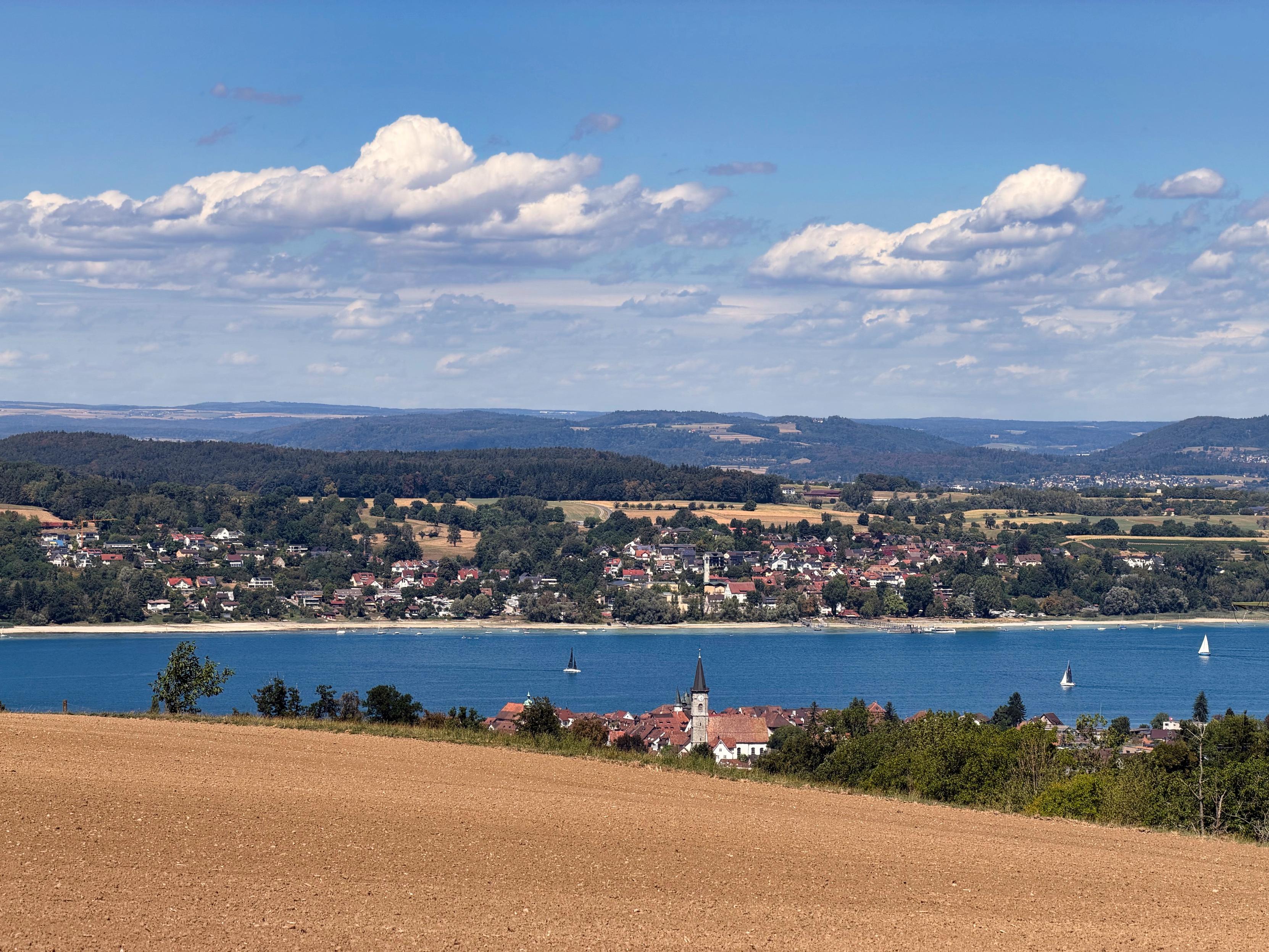 Eine malerische Landschaft mit Blick auf einen ruhigen See mit Segelbooten, umgeben von einem malerischen Dorf und sanften Hügeln unter einem teilweise bewölkten Himmel. Der Vordergrund enthält ein gepflügtes Feld.