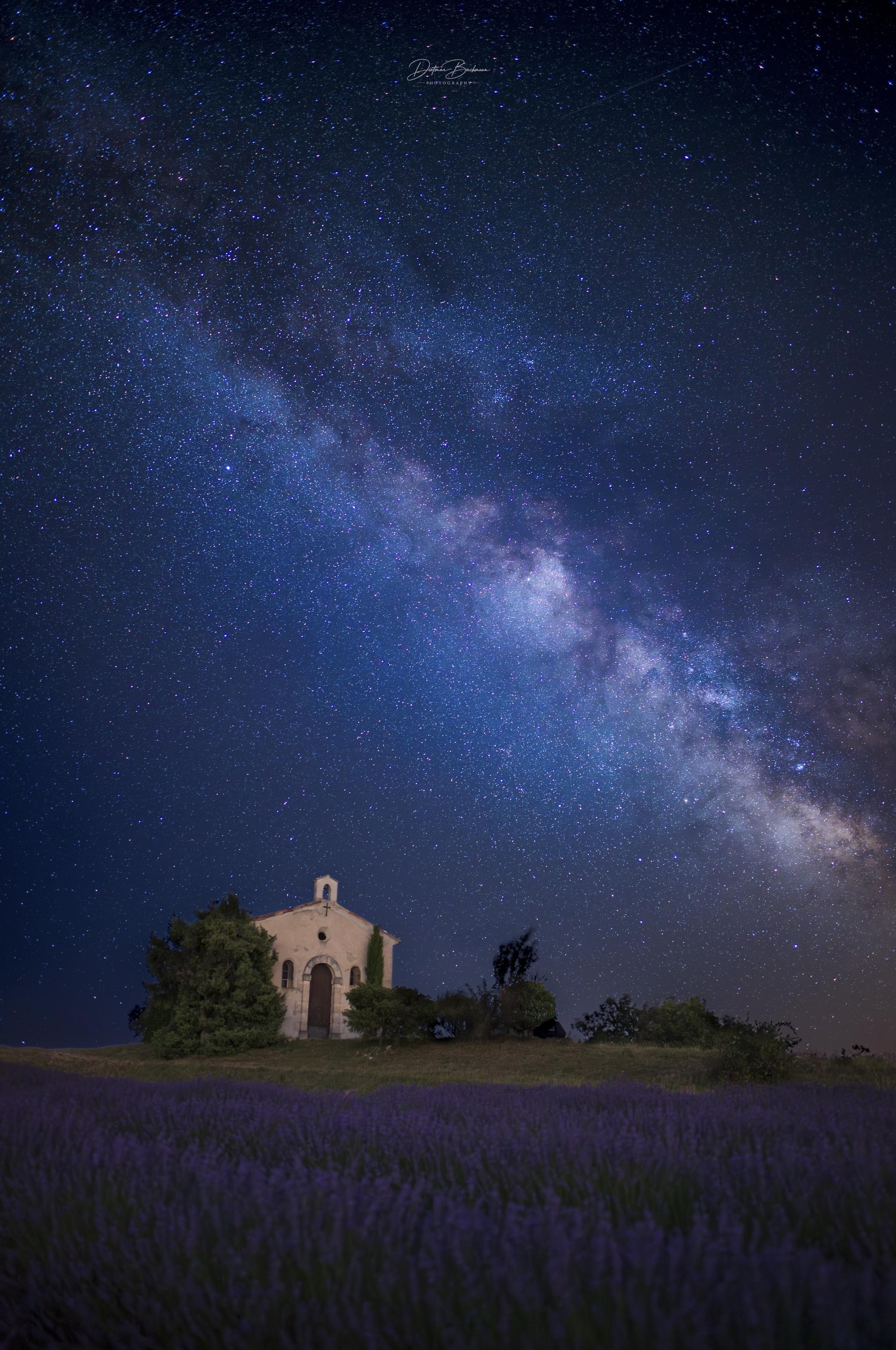 A serene night scene featuring a small chapel surrounded by vibrant lavender fields under a starry sky. The Milky Way is prominently visible, creating a stunning contrast against the dark backdrop.