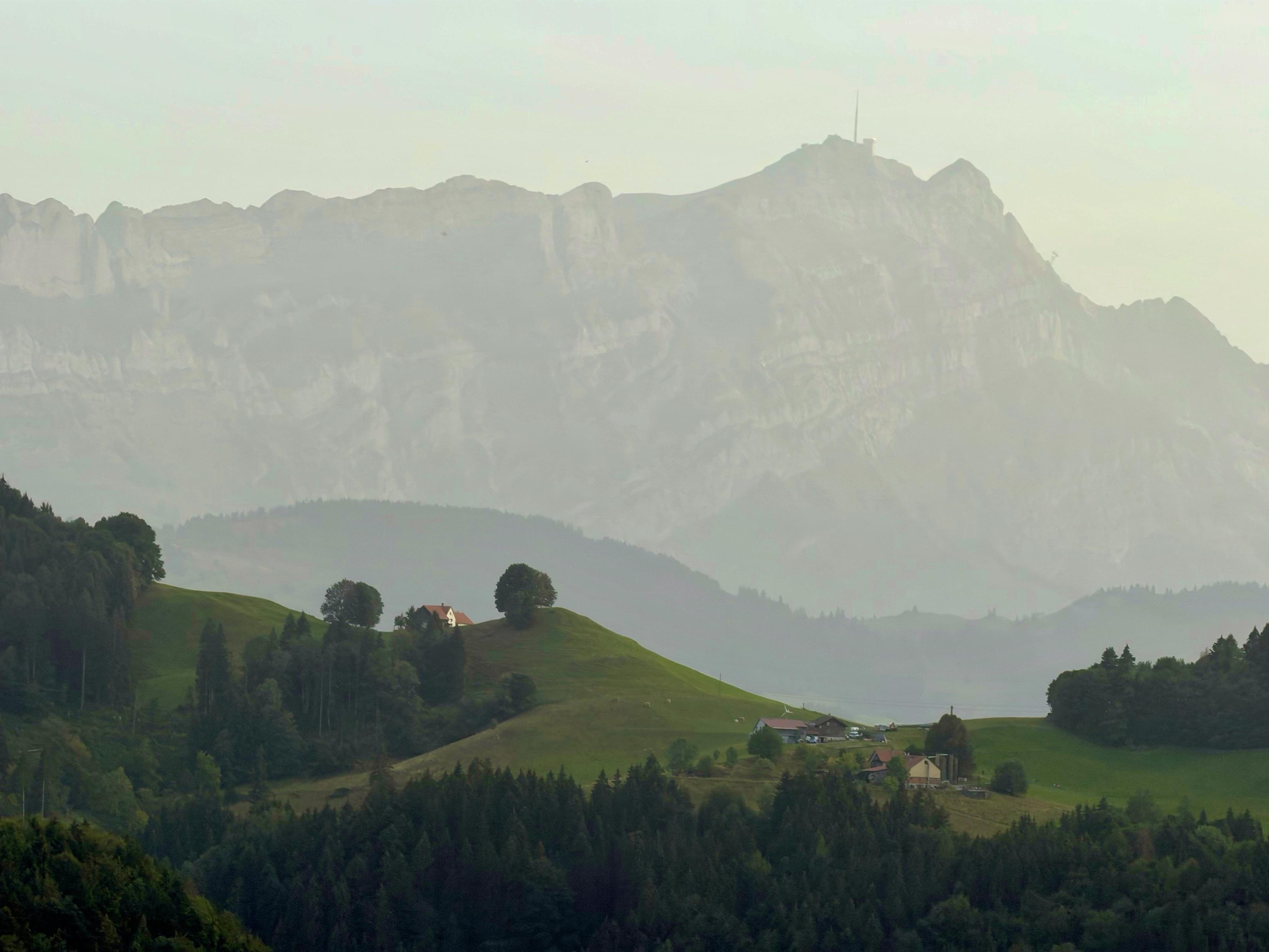 Eine ruhige Landschaft mit sanften Hügeln mit verstreuten Bäumen und kleinen Häusern, vor der Kulisse majestätischer Berge, die in Nebel gehüllt sind.