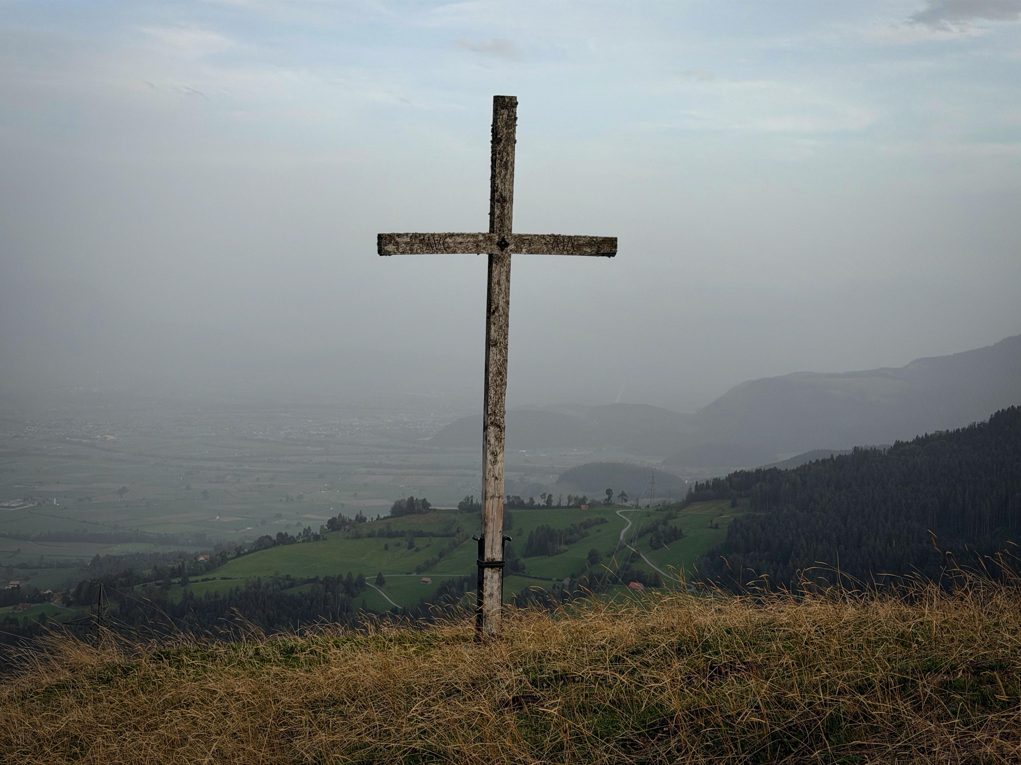 Ein verwittertes Holzkreuz steht auf einem Hügel mit Blick auf eine neblige Landschaft mit sanften Hügeln und Tälern. Die Szene zeigt Flecken von grünem Gras und verstreute Bäume unter einem bewölkten Himmel.
