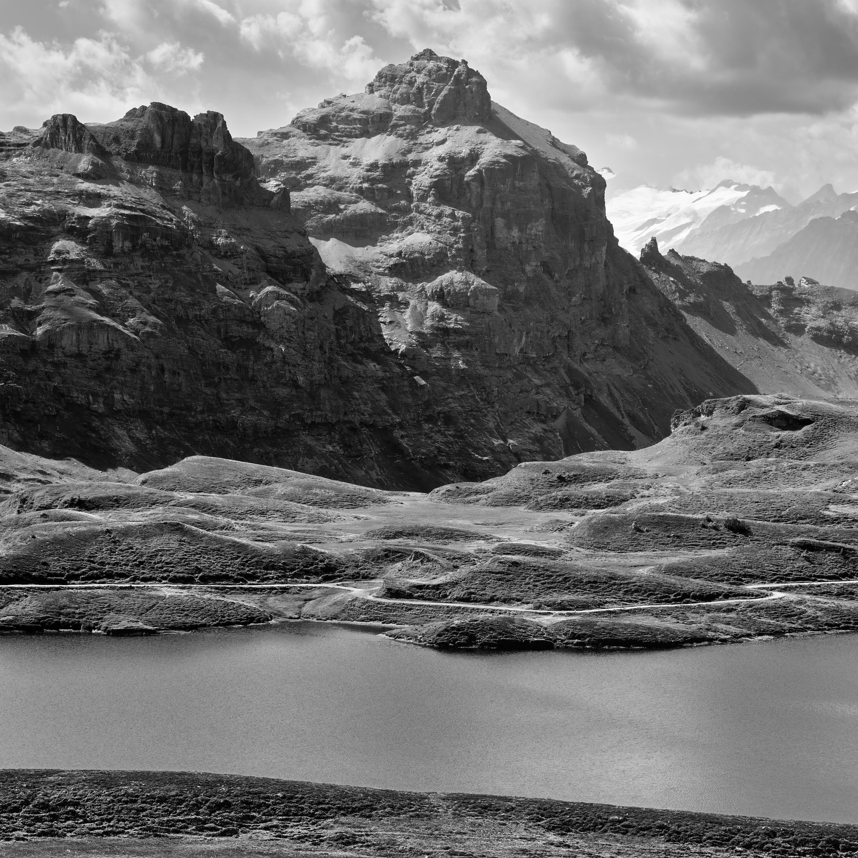 A dramatic black-and-white landscape featuring rugged mountains, rolling hills, and a tranquil lake in the foreground. The scene conveys a sense of vastness and natural beauty, with a cloudy sky above.