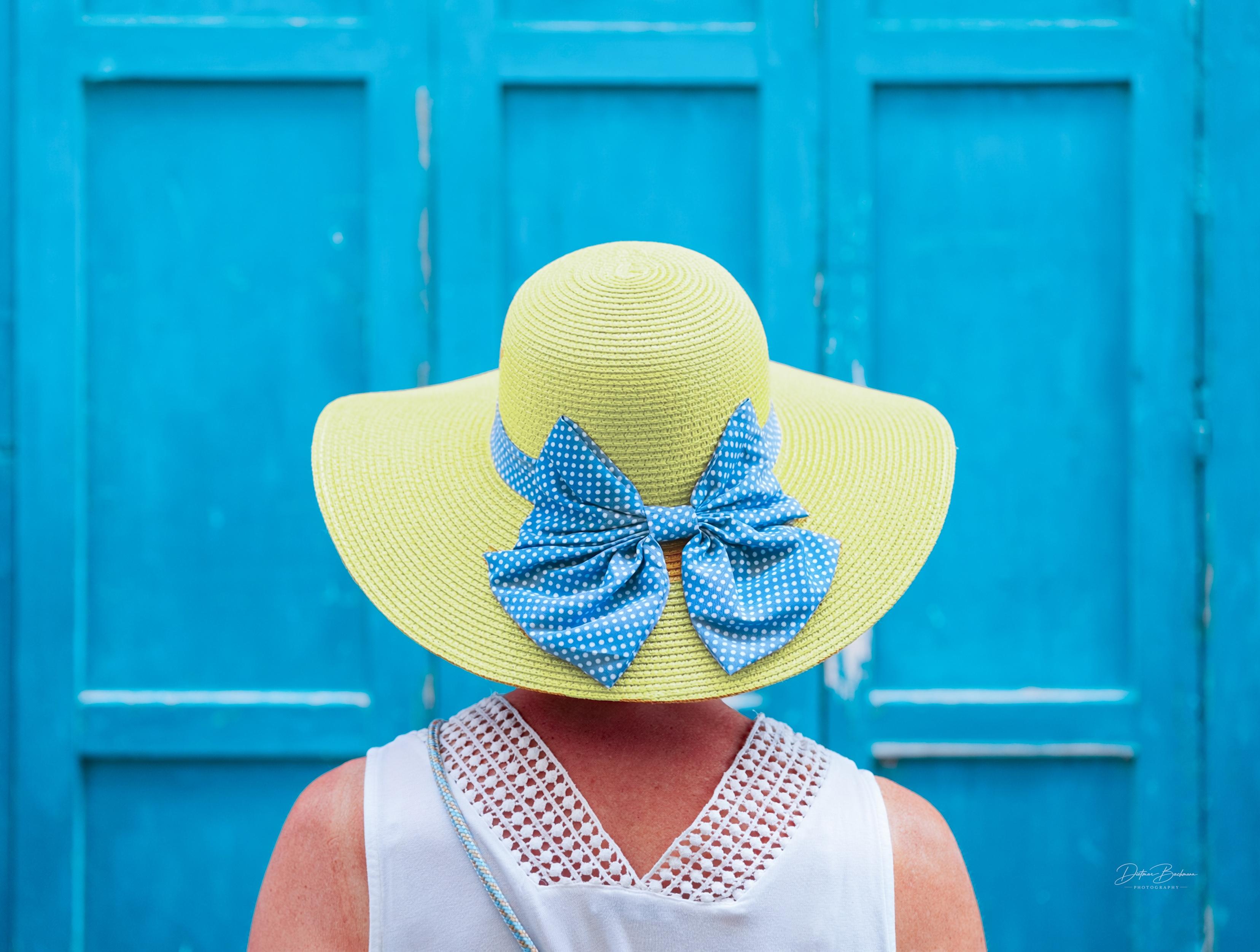 A person is seen from behind, wearing a wide-brimmed yellow straw hat adorned with blue polka dot bows. The background features a vibrant blue wall with wooden panels.