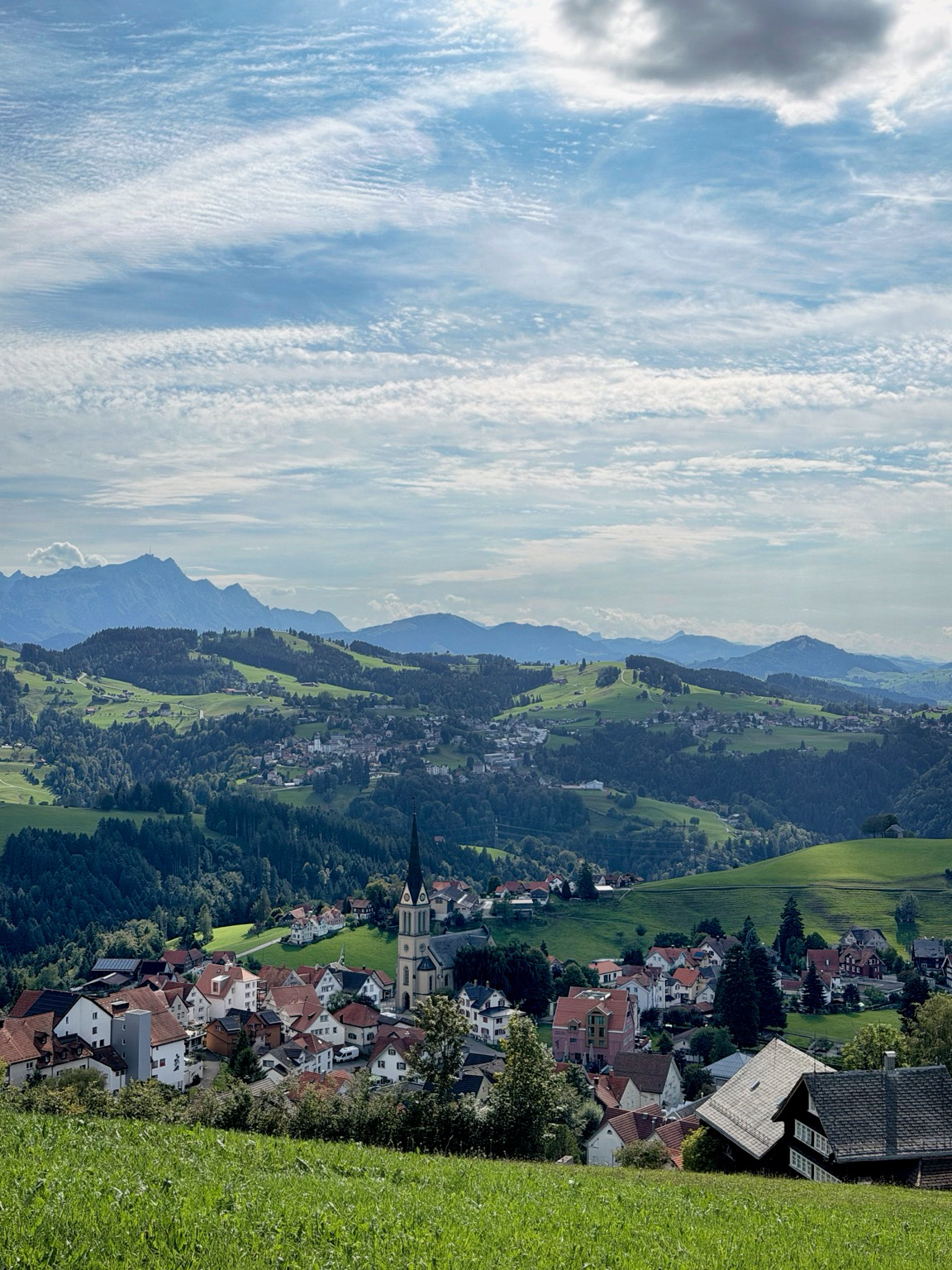 ein kleines dorf vor grünen, teils bewaldeten hügeln und einer bergkette. der himmel ist teilweise bedeckt