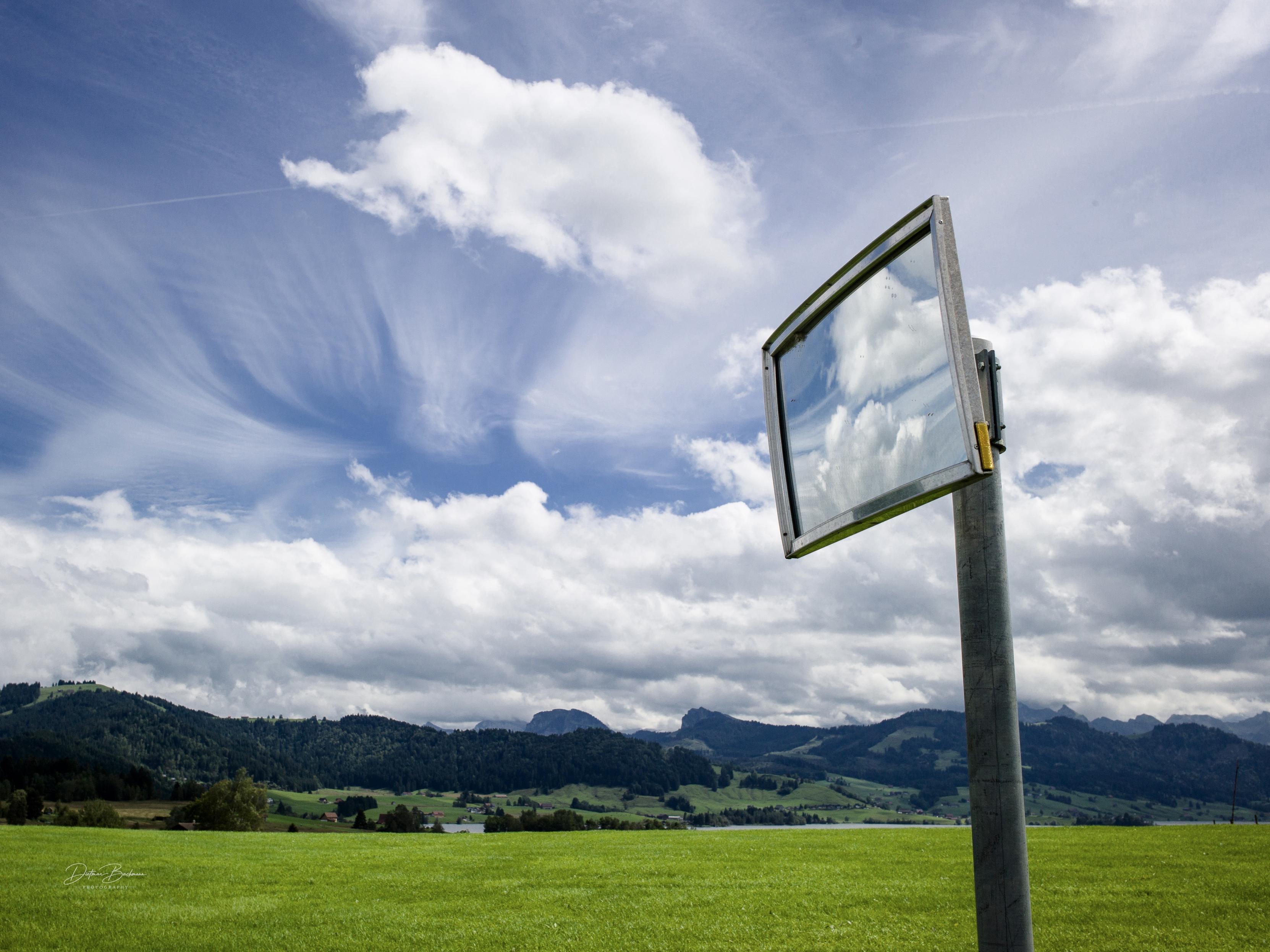 A reflective signpost stands in a green field under a partly cloudy blue sky, showcasing clouds and distant mountains in its reflection.