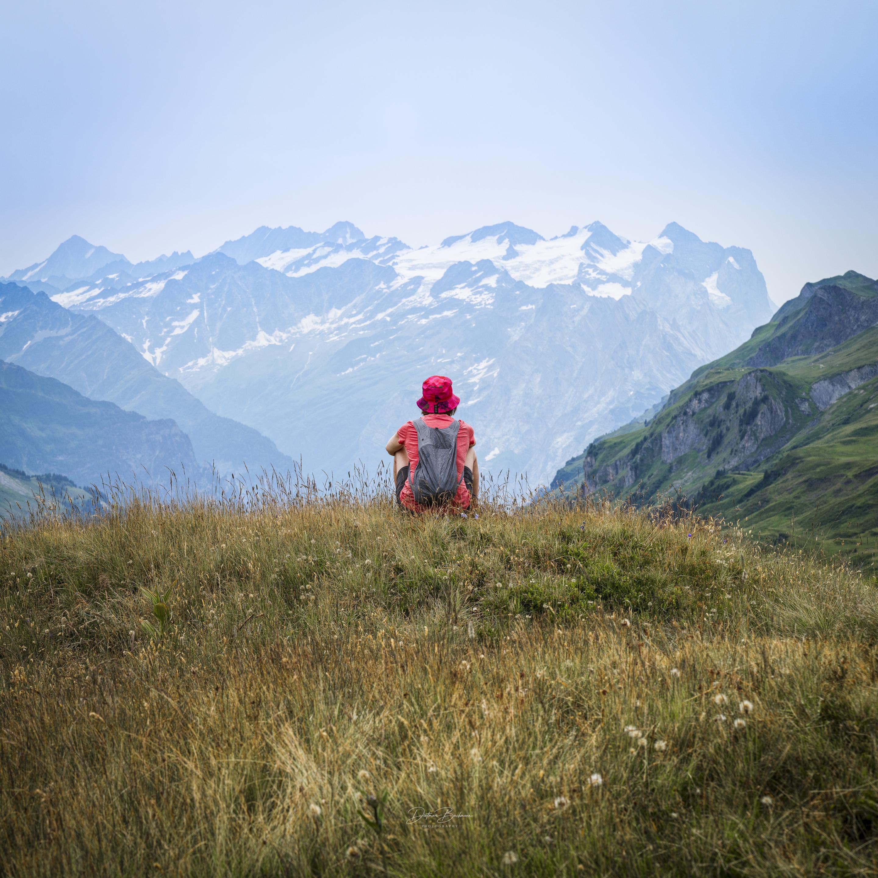 A person wearing a red hat and backpack sits on a grassy hill, facing a panoramic view of mountains and valleys in the background, under a cloudy sky.
