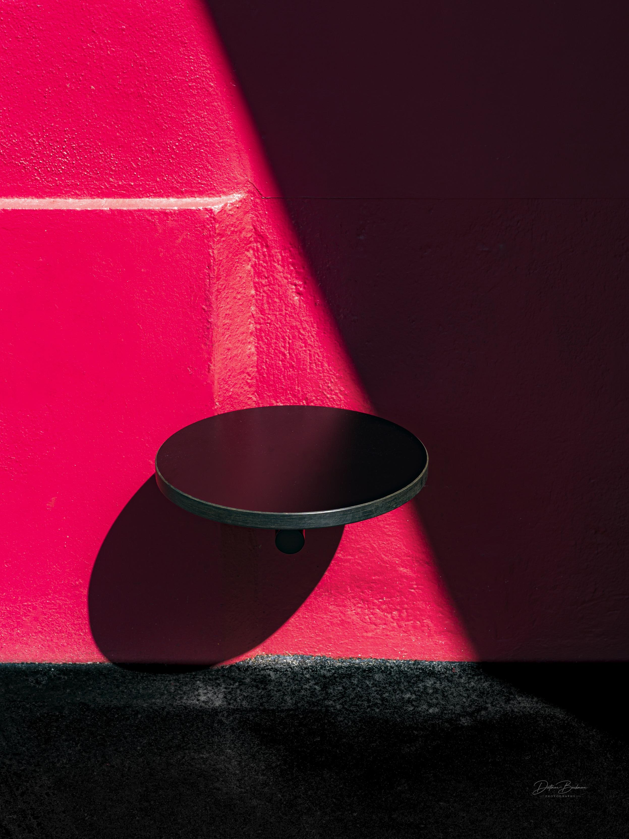 A black circular shelf is mounted on a bright pink wall, casting a shadow. The image features strong contrasts of color and light.