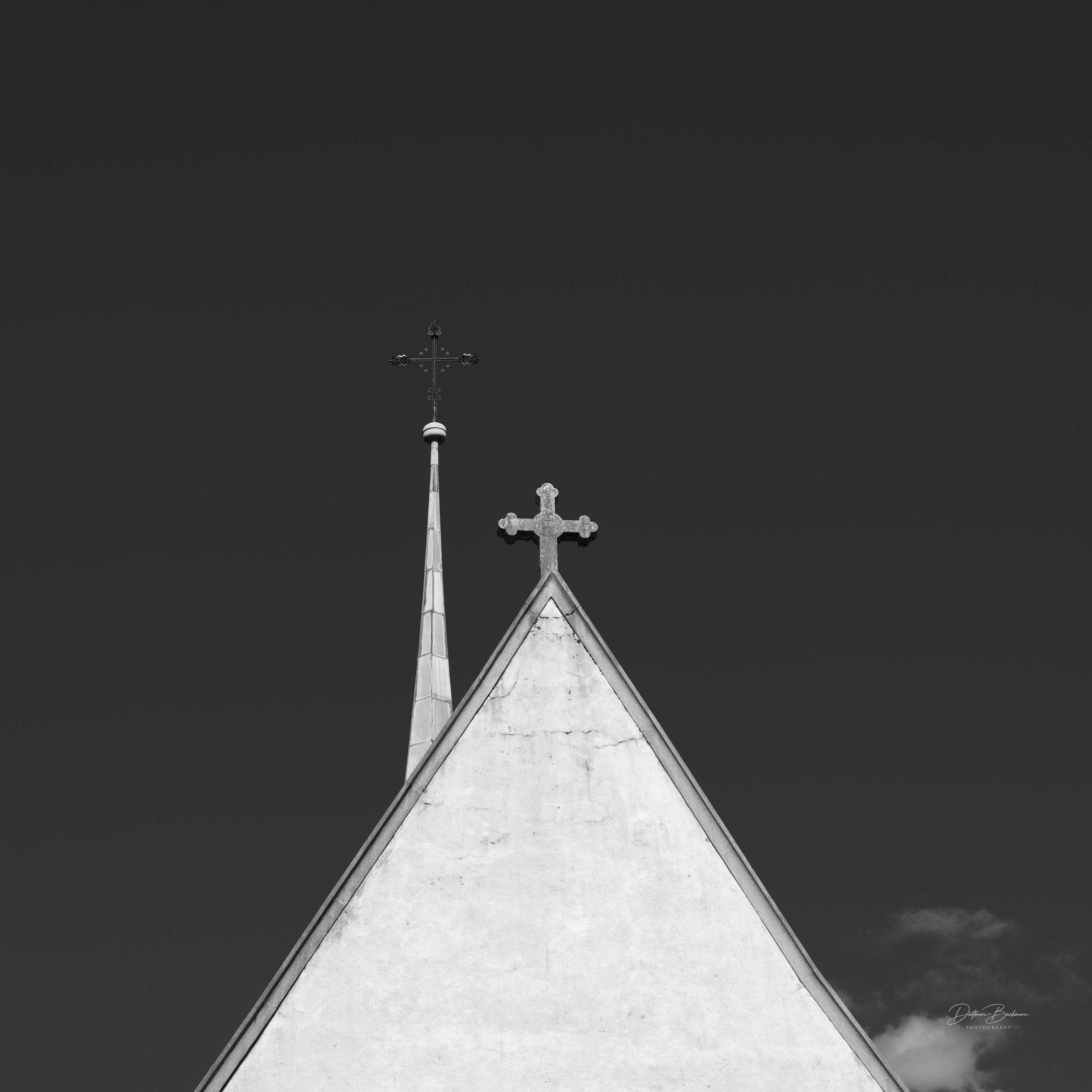 A close-up view of church architecture featuring a peaked roof and two crosses, one on a spire and another at the apex. The image is presented in black and white against a plain dark background.