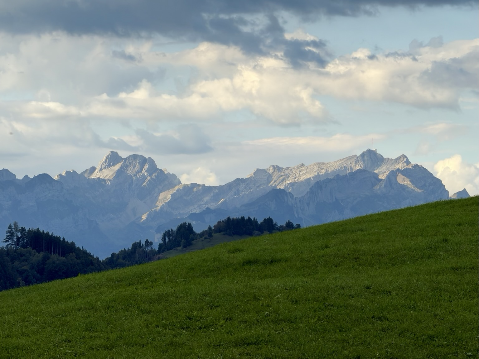 eine grüne wiese, dahinter ein bergmassiv, darüber wolken