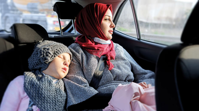 Eine Frau sitzt auf der Rückbank in einem Auto und neben ihr ein Kind welches schläft
