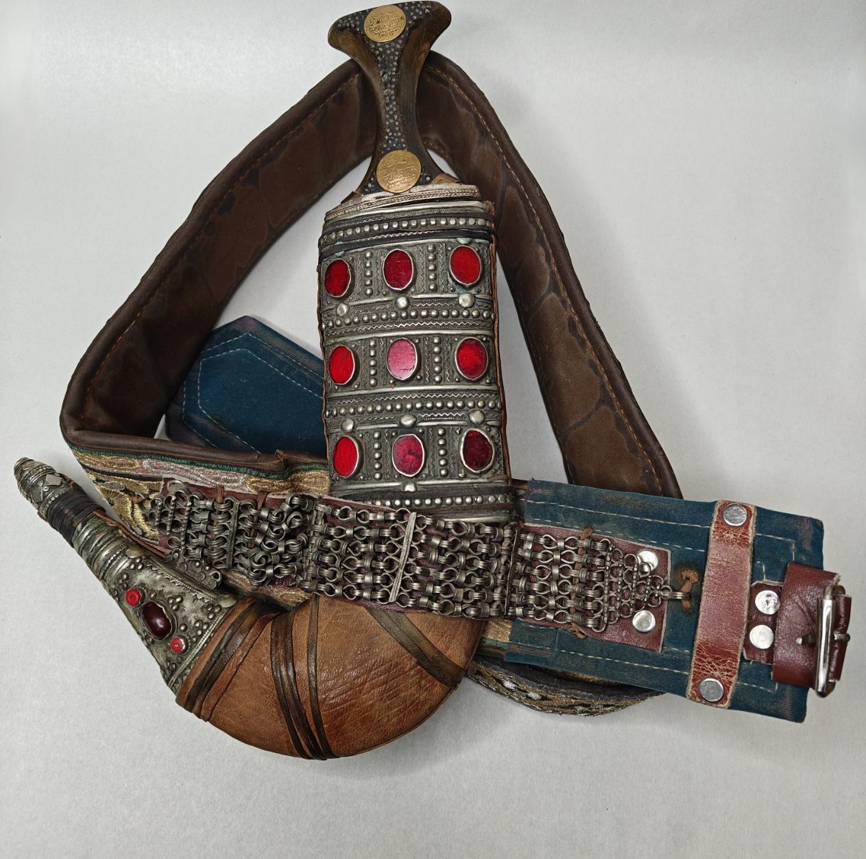  Jambiya dagger  with belt has red glass beads inlaid in old silver. Dagger is in a leather case that is attached to a wide belt that has green felt at the ends with silver and gold thread throughout giving a design 