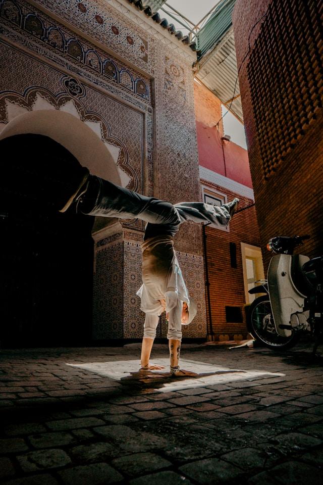 Photo. Person doing a handstand, legs split to run parallel with the ground and perpendicular to the torso. They appear to be wearing wide-leg jeans, white long-sleave shirt with the sleeves hiked up, and a head covering. The person does this at the intersection of buildings. Each structure has a beautiful design. One with an arch and geometric tiling, the other made with bricks. Lighting comes in dramatically from above