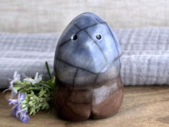a vaguely dome-shaped nature spirit ceramic sculpture with a gently pointed head and four stubby little legs. it's glazed in a bottom-to-top gradient of brown to blue and has a gentle face with two round eyes and a stubby nose. it's standing on a piece of chestnut wood with grey muslin and natural linen behind. next to it on the left is flowering rosemary in blue-violet and white (with green leaves).