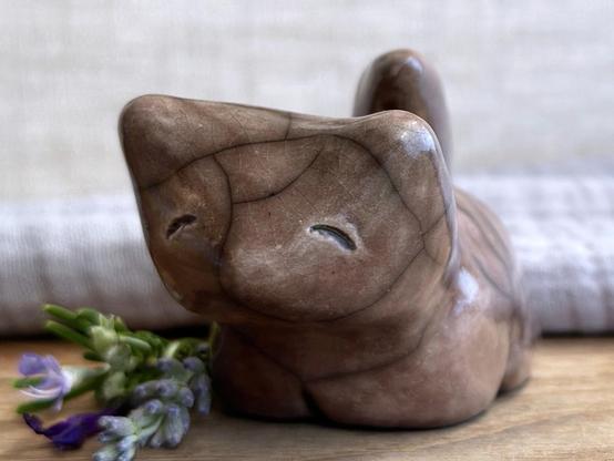 A sitting raku ceramic kitsune fox sculpture glazed in warm brown with hints of mauve. It has a very kind, gentle face, and its tail is curled up its back. It has four stubby legs and is standing on a piece of chestnut wood with grey muslin and natural linen behind. Next to it on the left are some lavender, an indigo-violet rosemary flower, and a dark violet autumn-sage flower.