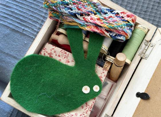 on a blue, woven-cotton sofa cover, a smallish white wooden box with hinges at the back. the lid is open, and the box is filled with various sewing bits: spools of thread in lots of colours, a few small scraps of fabric, a woven sort of braid lof thread in all colours of the rainbow and more, a wooden needle case, a wooden felting-needle holder, and a green felt rabbit shape with eyes that look like fried eggs. as an aside, i got the needle case many years ago at a re-enactment (when i was involved with such things) and it's one of my prized possessions.