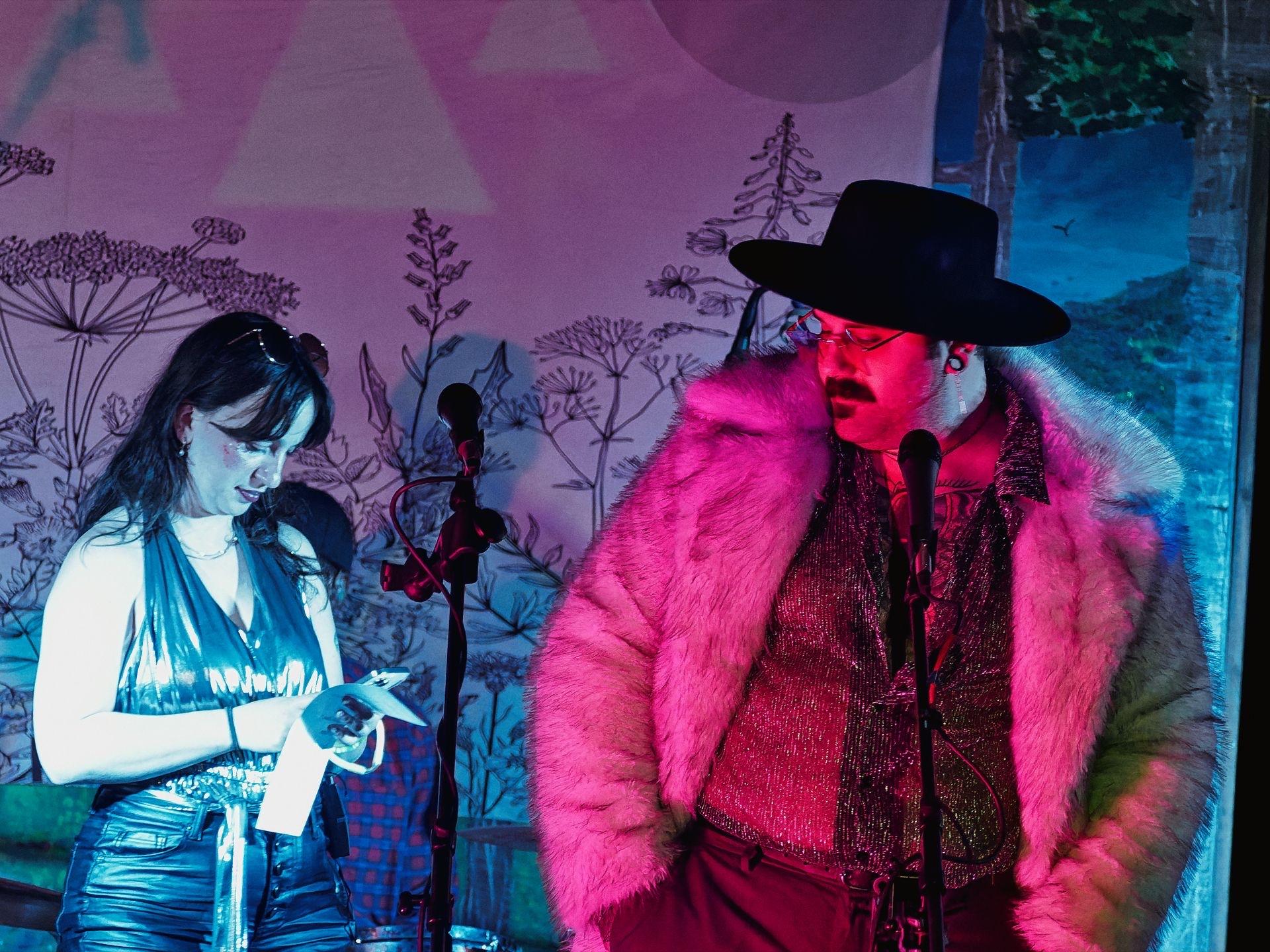 Two performers stand at microphone stands against a floral illustrated backdrop bathed in pink and blue stage lighting — on the left, a woman in a silver halter top and metallic jeans glances down at her phone, and on the right, a person in a wide-brimmed black hat, red glasses, a pink fur coat, and a glittery burgundy top leans slightly forward toward the mic.